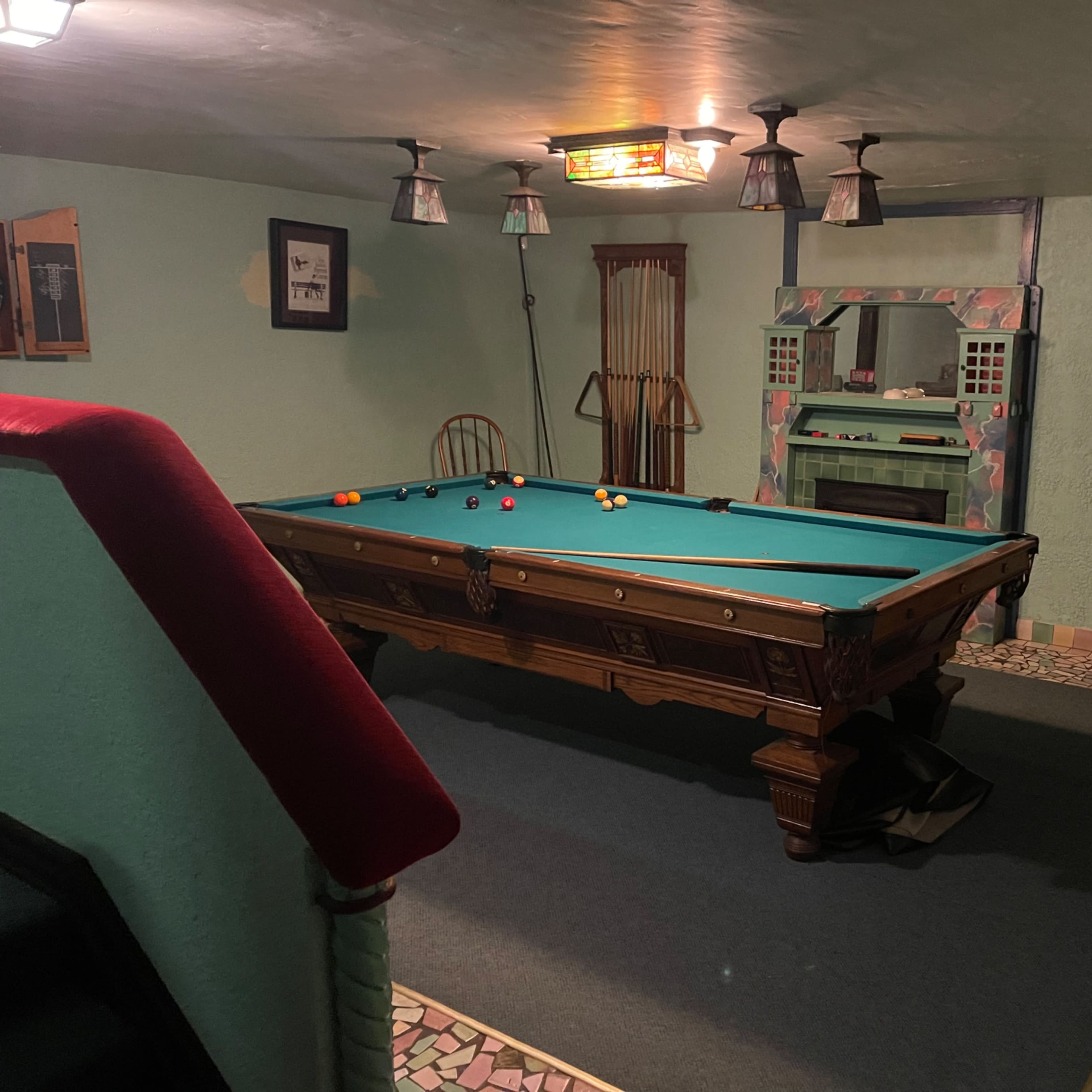 A basement game room featuring a pool table surrounded by several wooden cues, with a mirrored wall and mosaic tiled flooring.