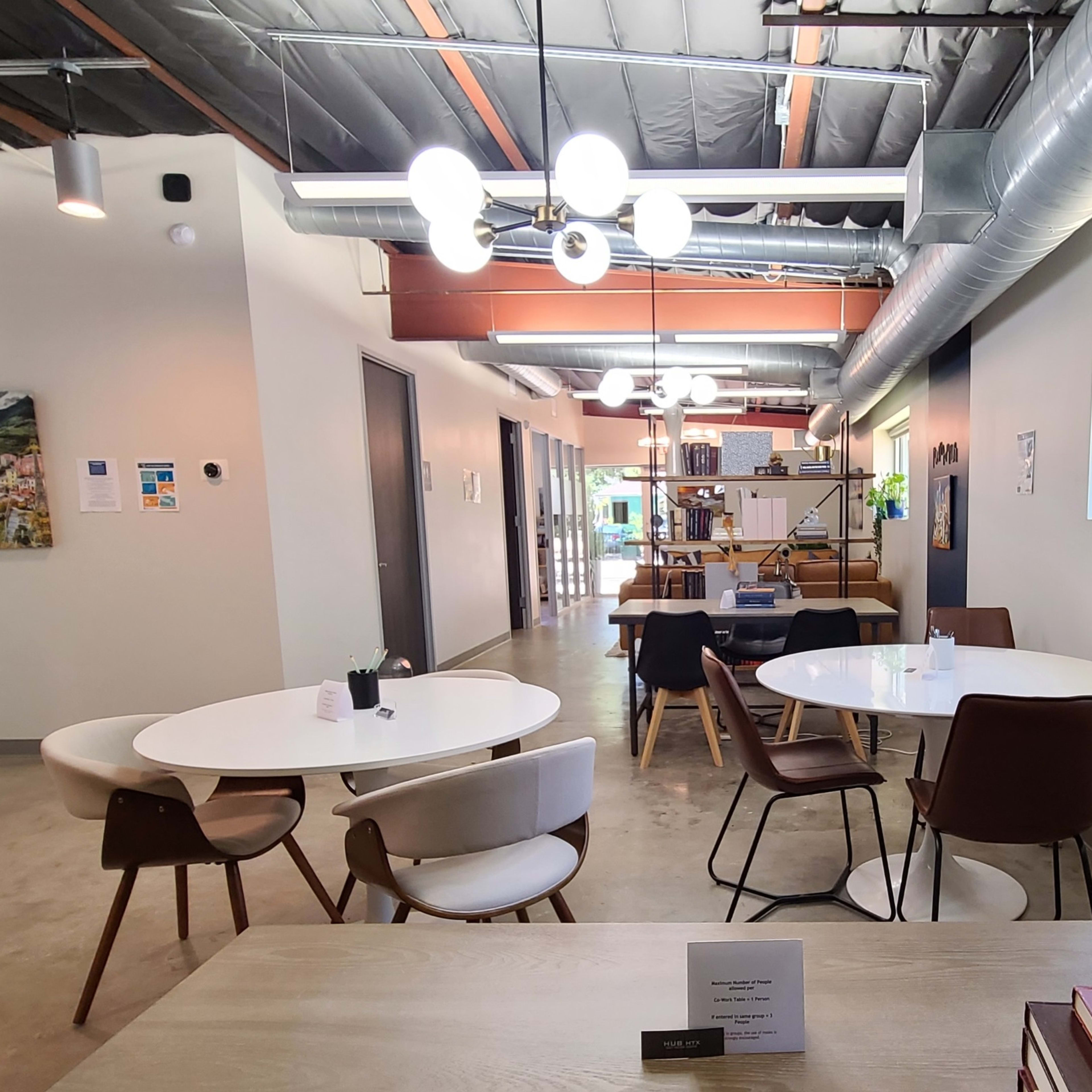 A modern co-working space with several white and wooden tables surrounded by chairs, and plants placed in the windows.