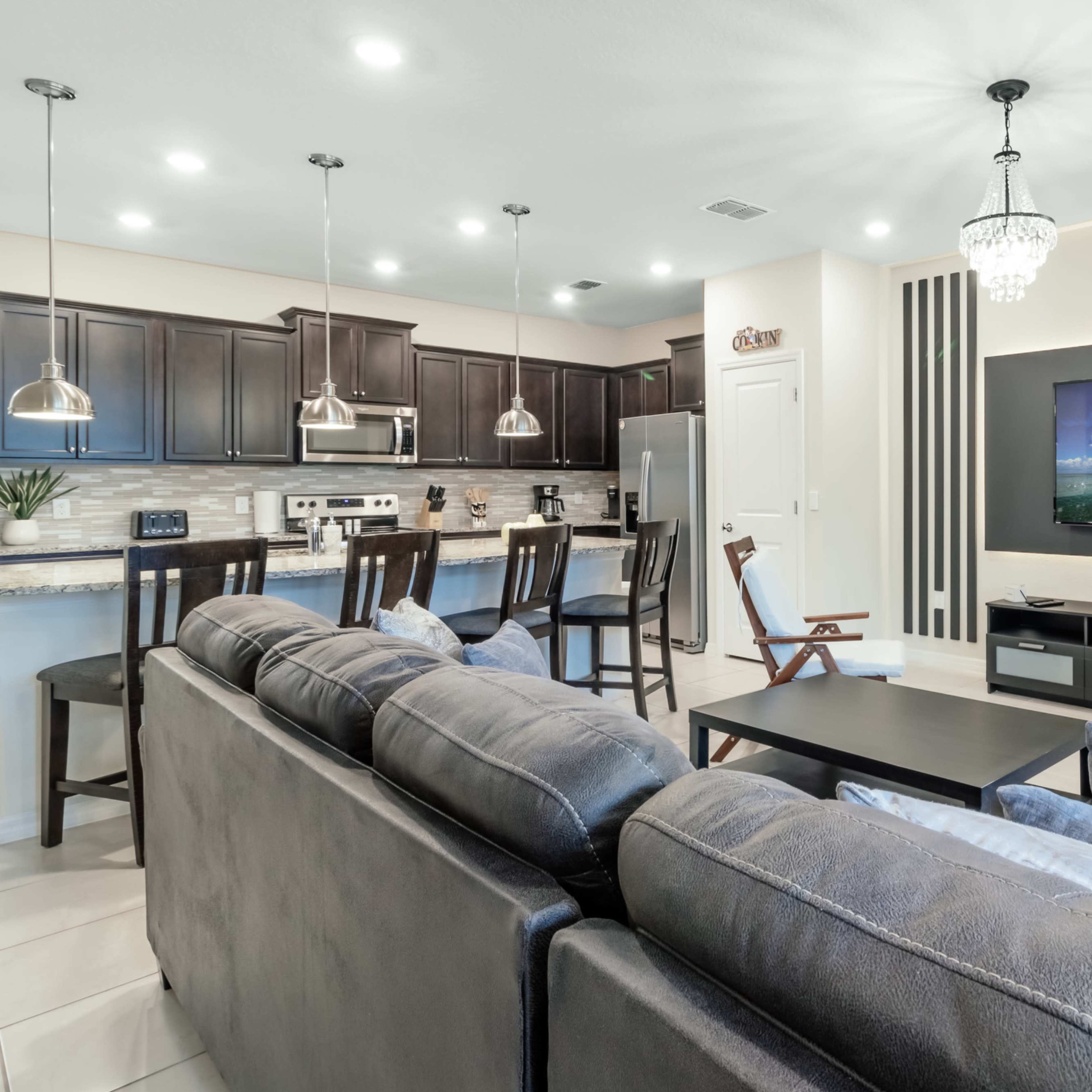 A modern living room with a gray sectional sofa, a black coffee table, a large wall-mounted television, and an open kitchen area with dark cabinets and stainless steel appliances.