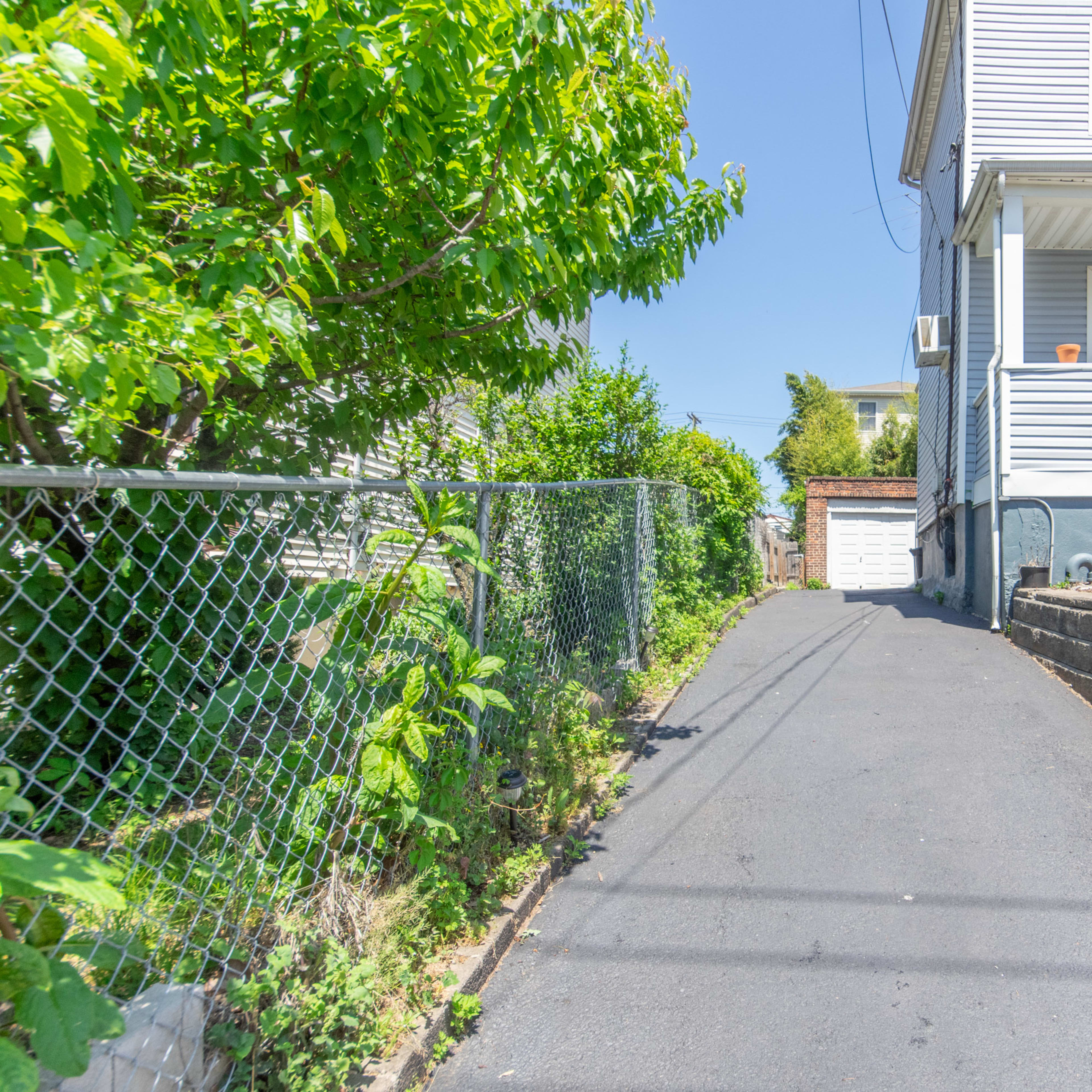 A narrow asphalt driveway lined with greenery leads to a white garage at the back of a residential property.