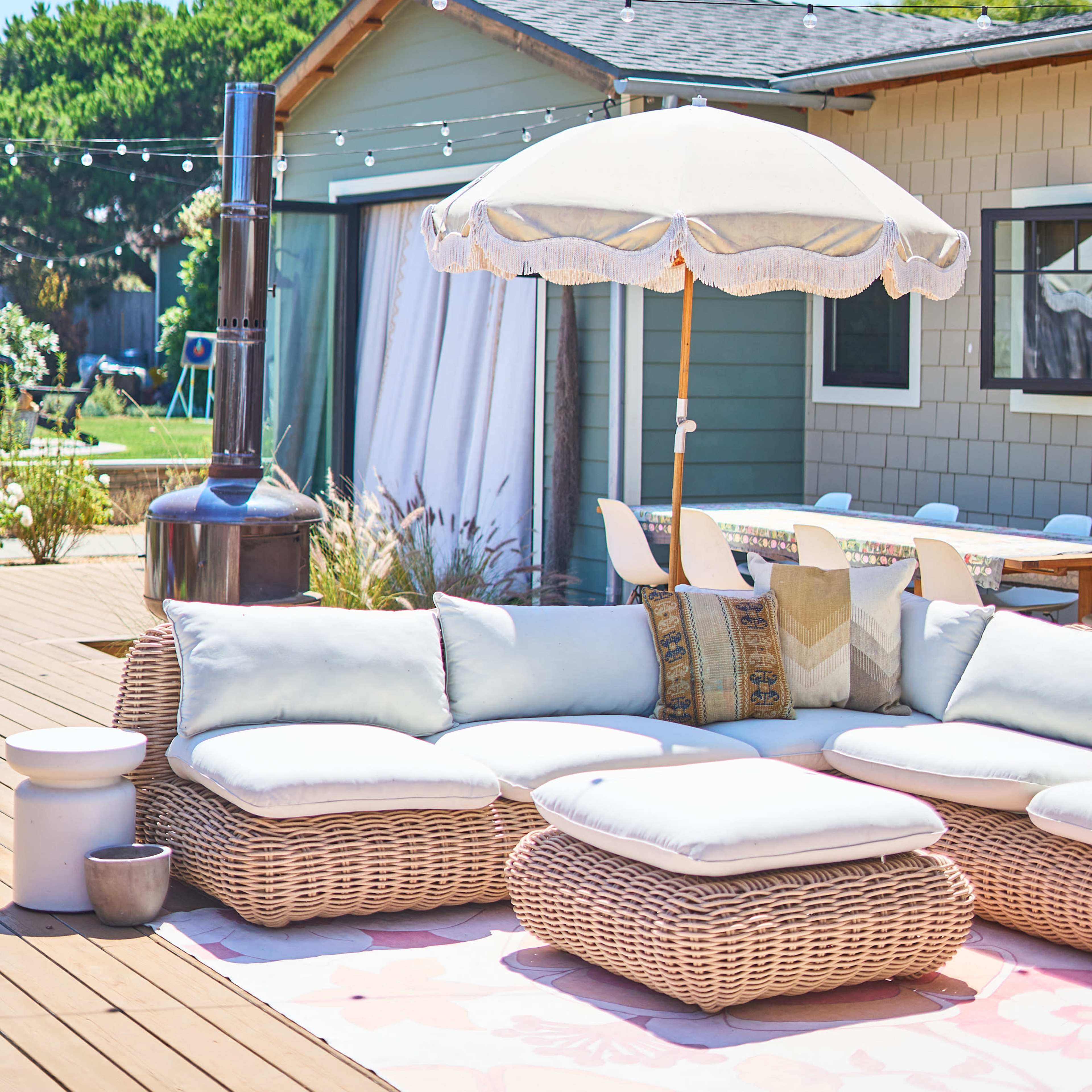 A patio area features a large wicker sectional sofa with cushions, a decorative umbrella, and a backdrop of a house and outdoor seating.