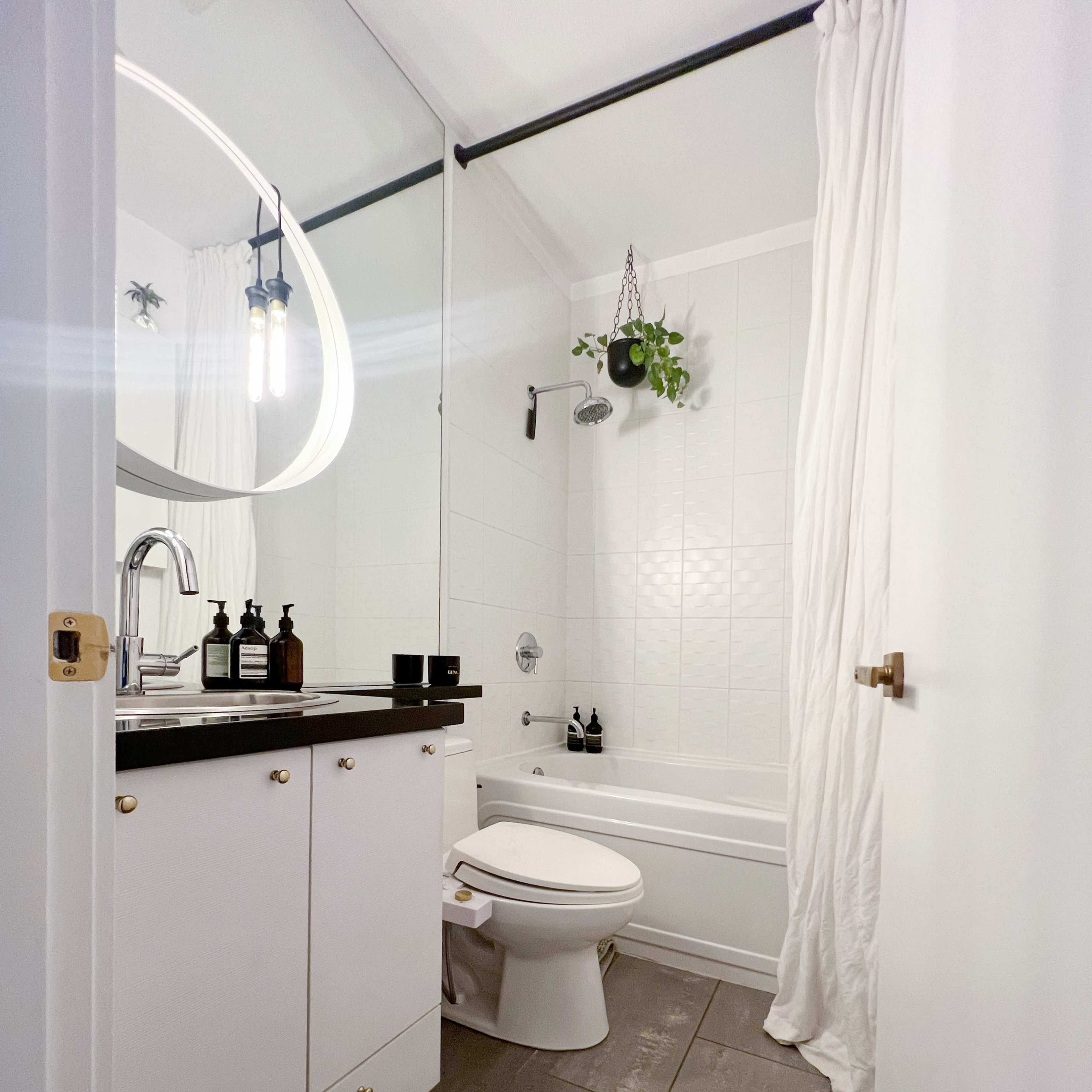 A modern bathroom with a bathtub, a toilet, a sink with dark countertop, and white cabinets, featuring a mirror and a hanging plant.
