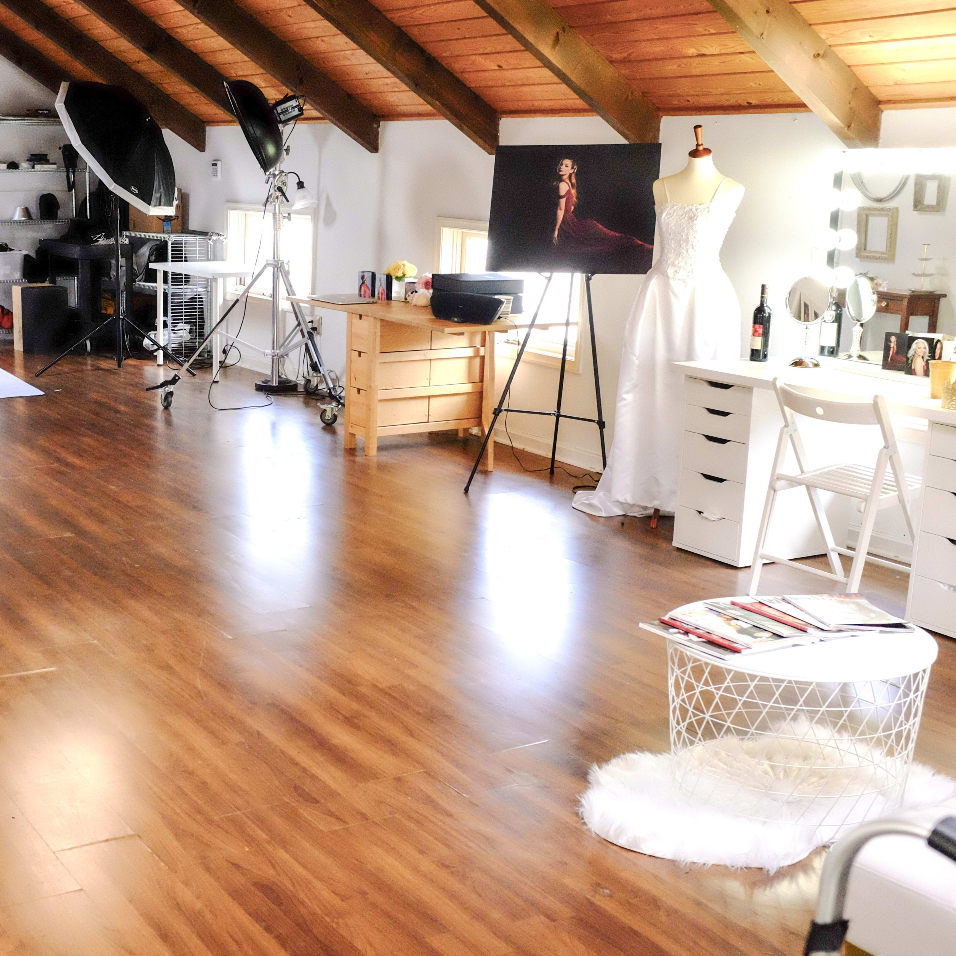 A well-lit photo studio with wooden flooring, equipped with various photography equipment and a garment rack displaying a white dress.