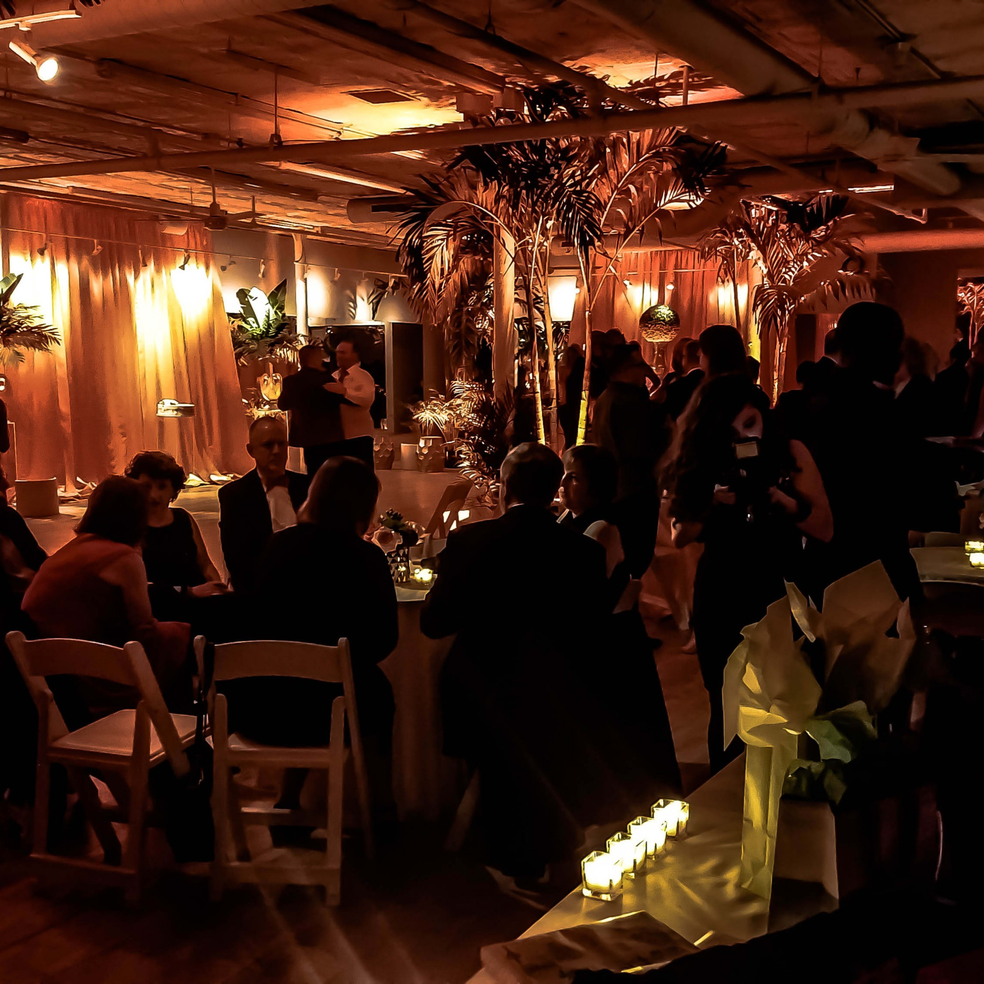 A dimly lit event space is decorated with tropical plants and candles, with guests seated at tables and mingling.