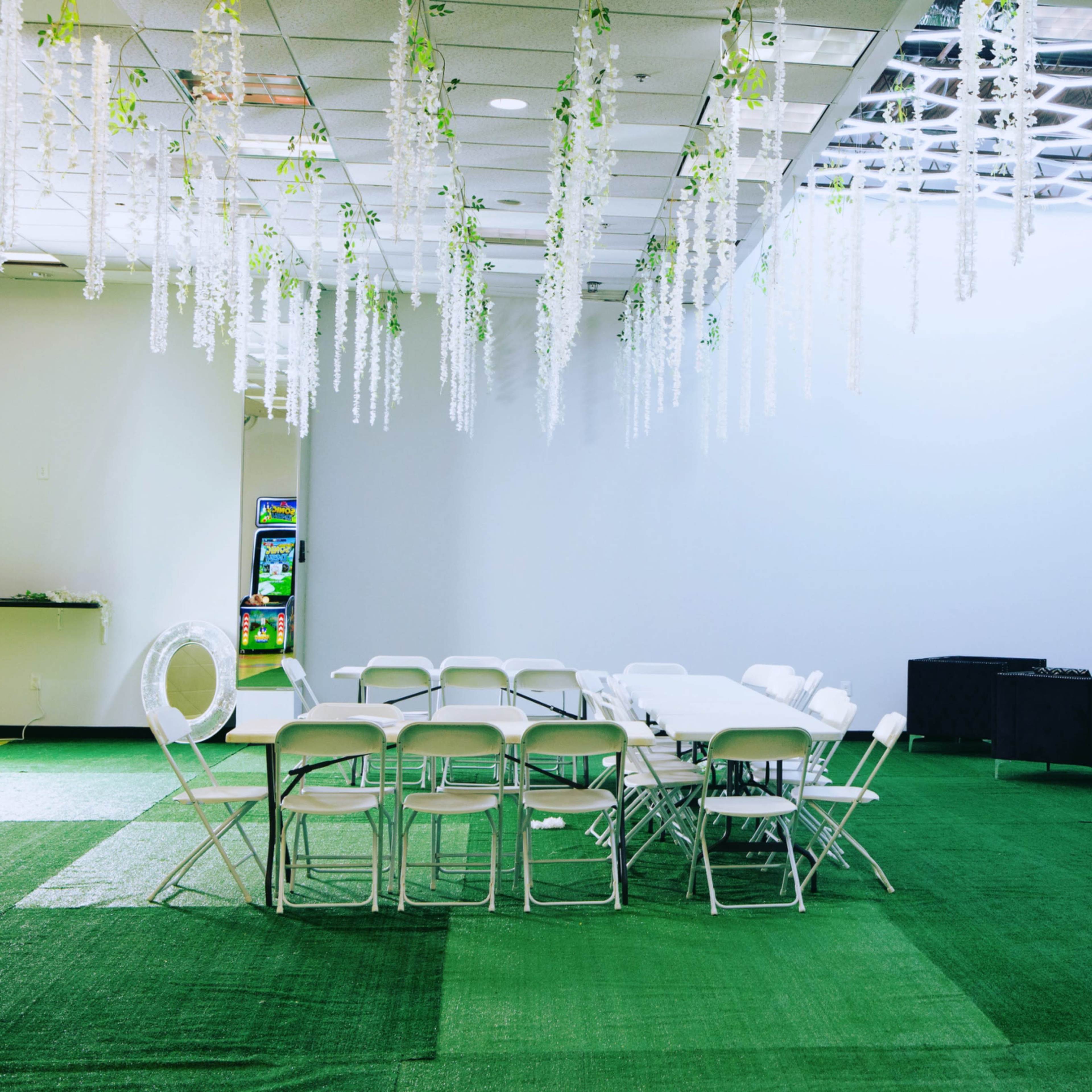 A room with green carpet features a circular table arrangement with white folding chairs and decorative hanging plants.
