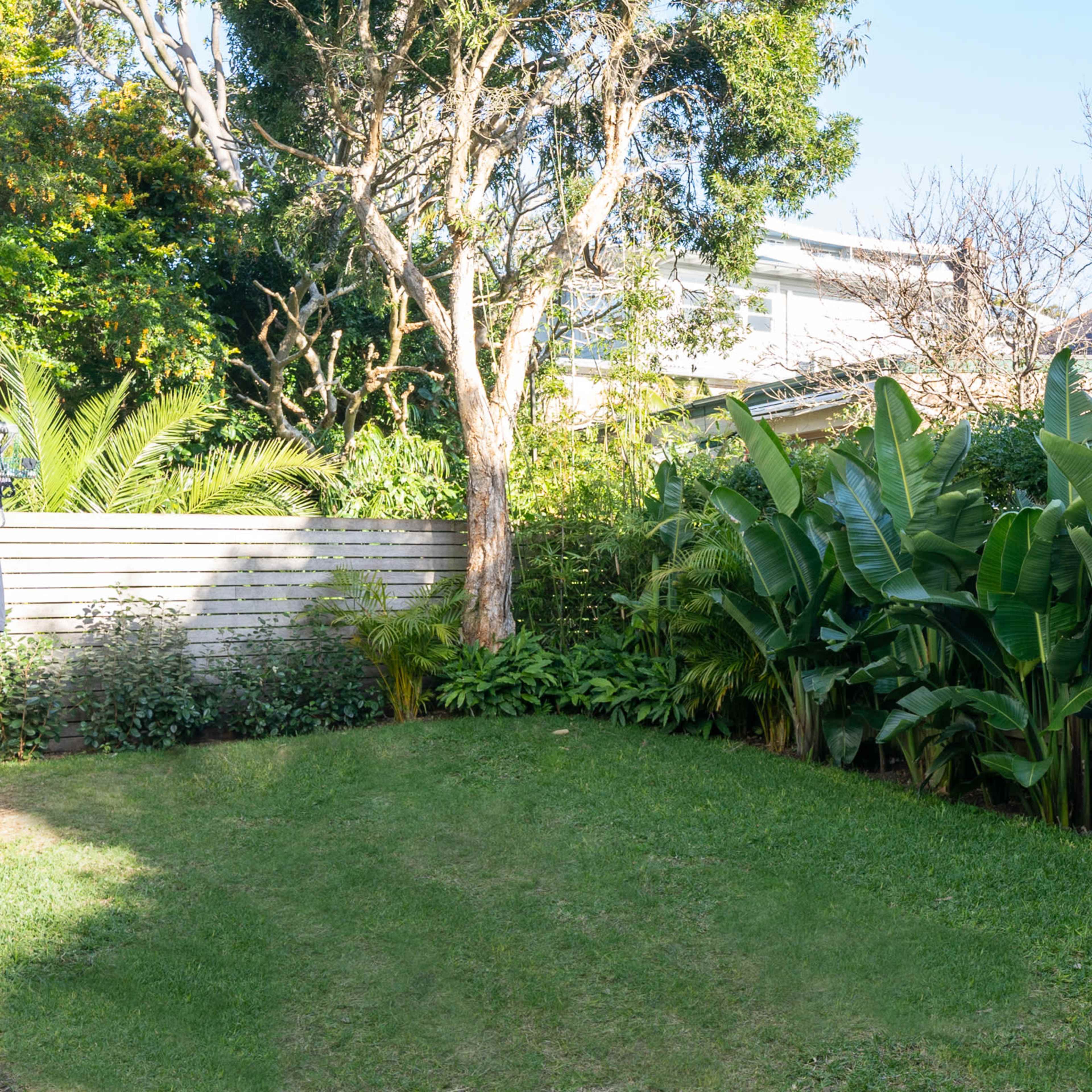 A neatly maintained backyard featuring lush green plants and trees, bordered by a wooden fence and neighboring houses in the background.