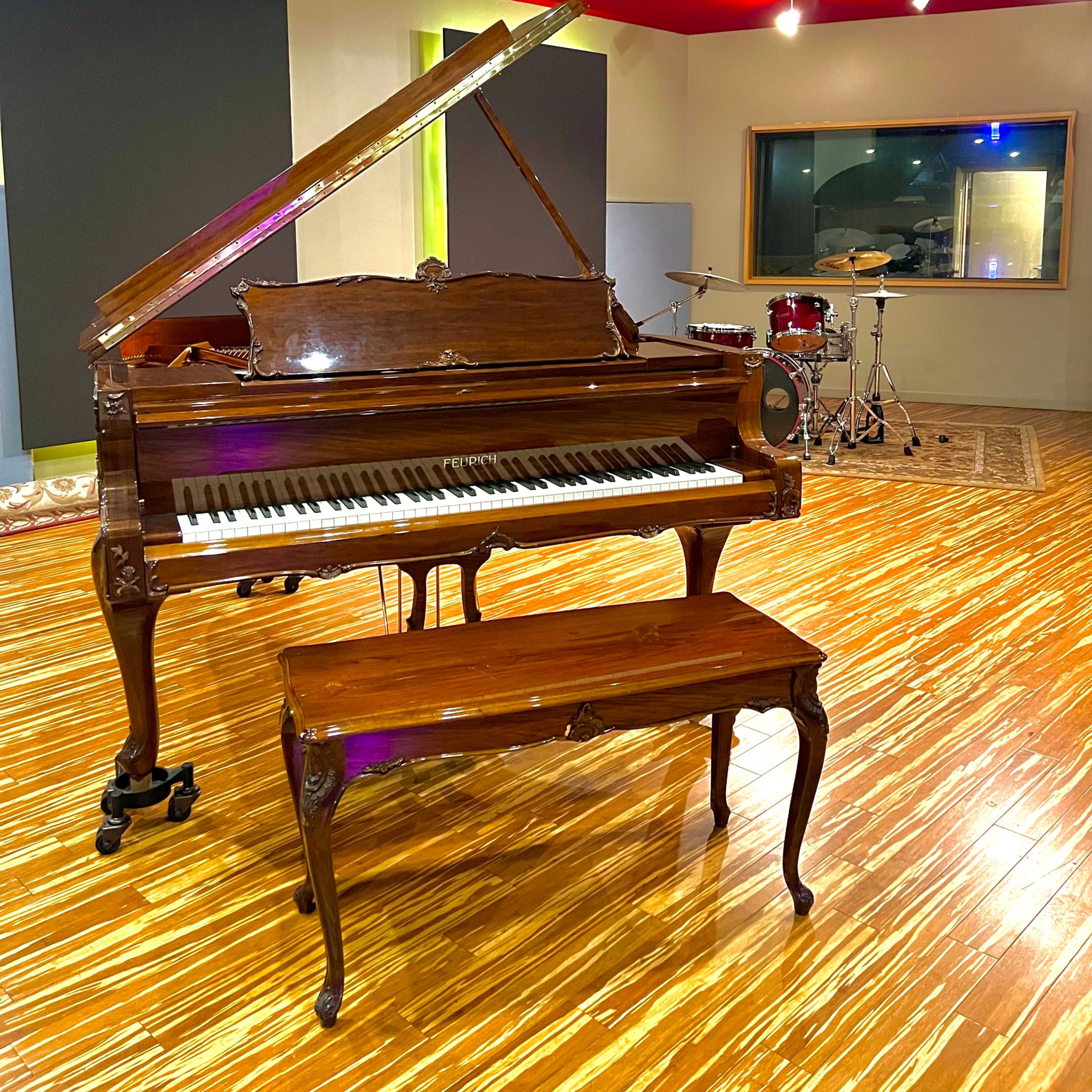 A grand piano with an opened lid and an accompanying bench sits on a polished wooden floor in a music studio, with a set of drums visible in the background.