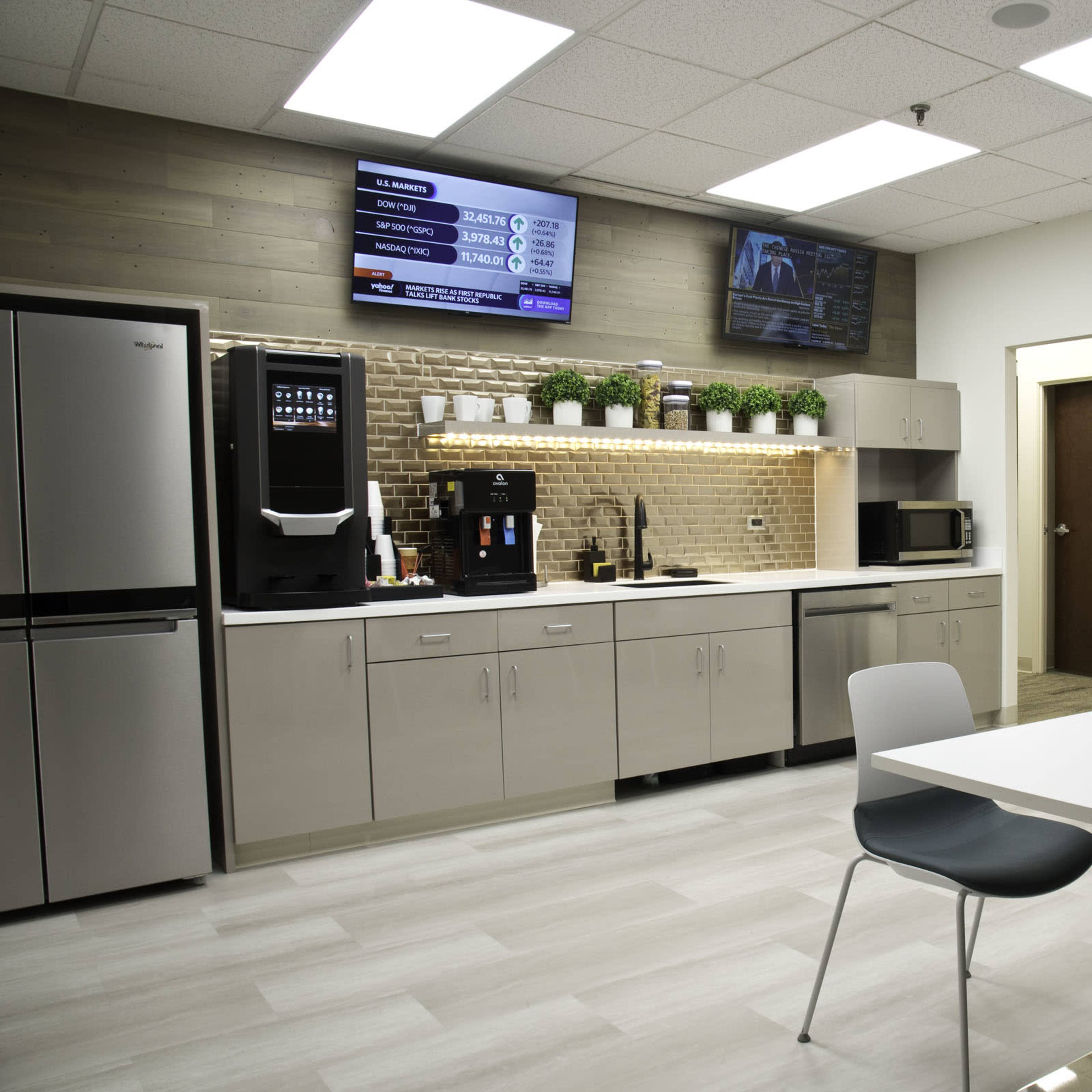 A modern kitchen area with stainless steel appliances, a countertop, and a seating area, featuring two televisions on the wall.