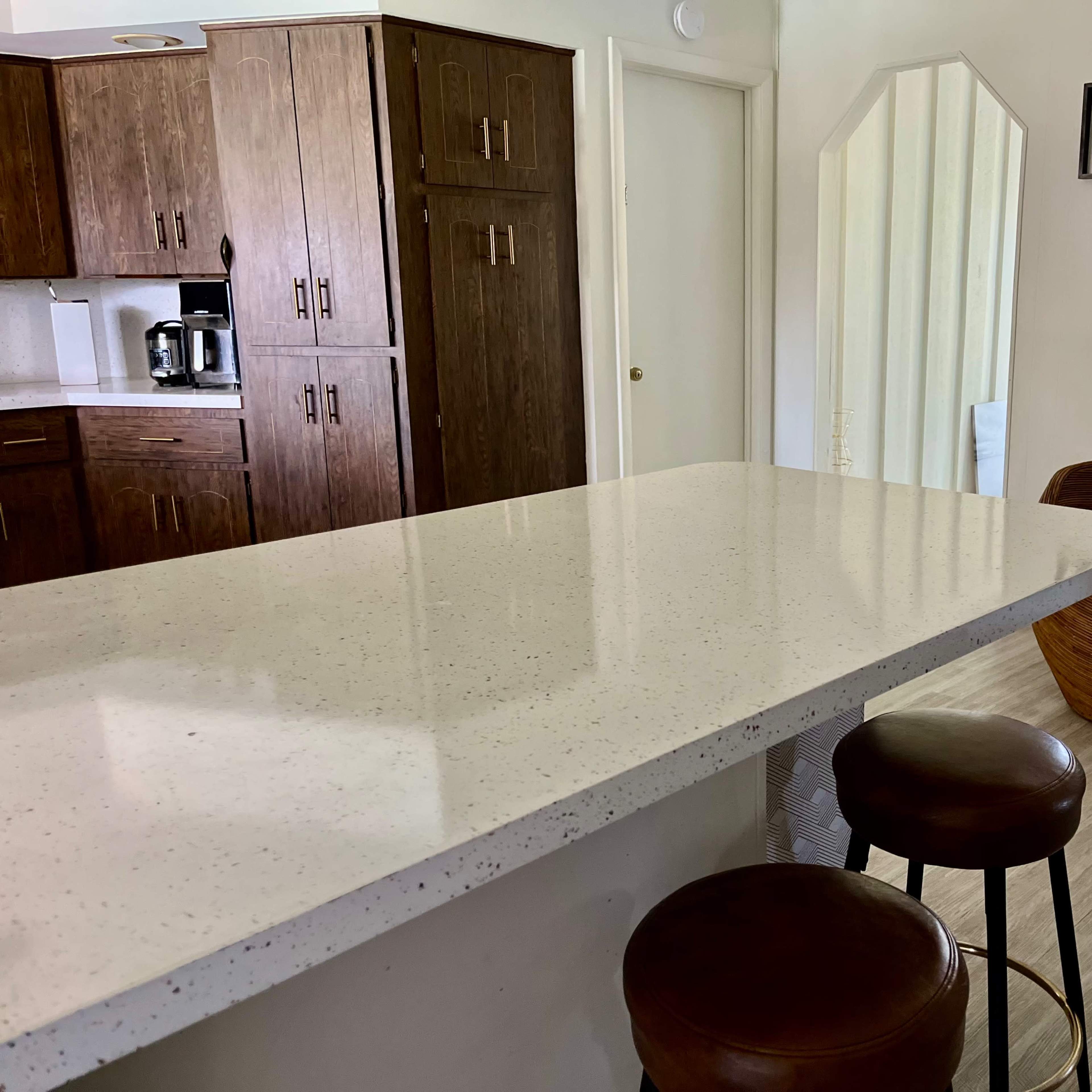 A kitchen area with a white countertop and two bar stools, adjacent to a cozy seating area with a mirror and decor.
