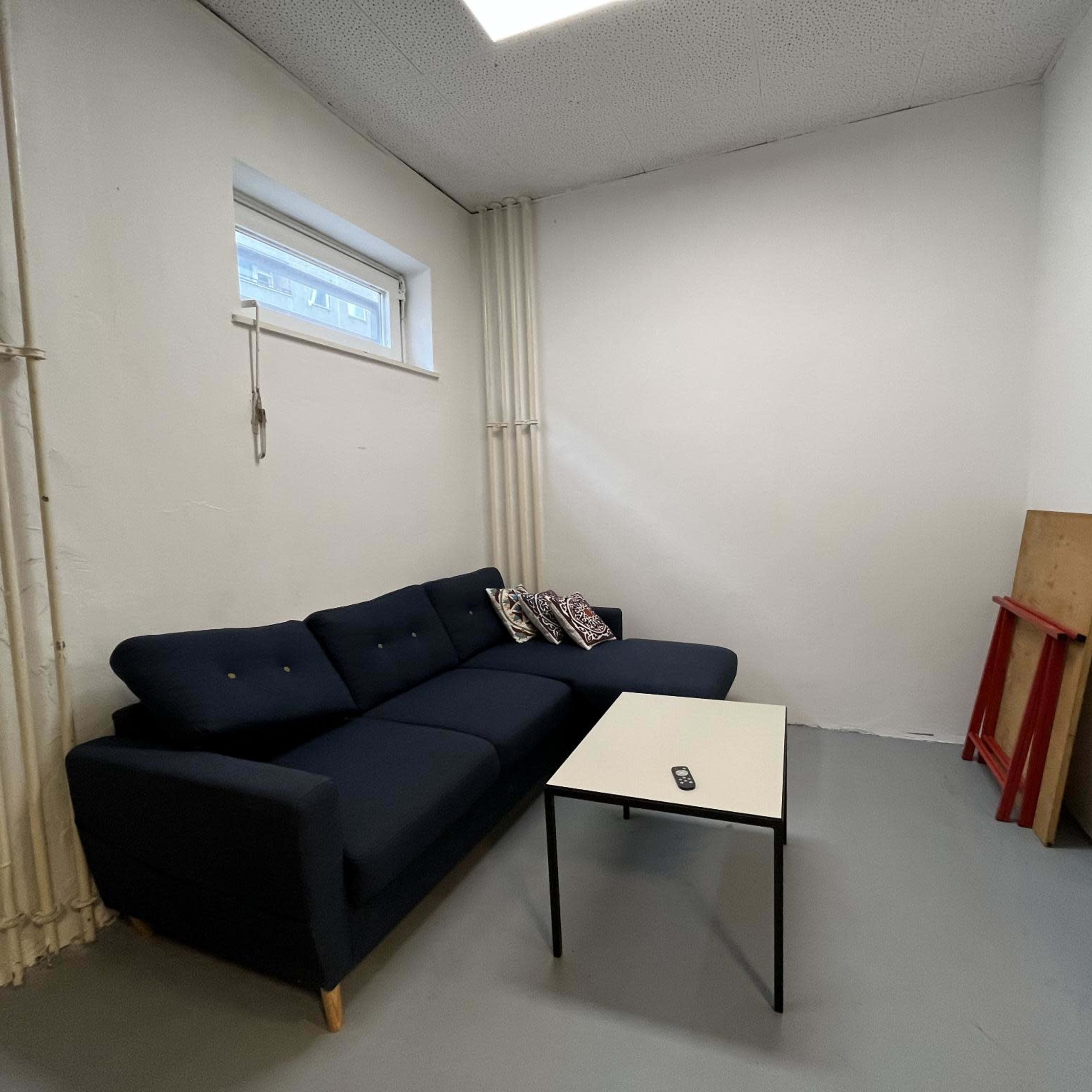 A small room features a dark sofa with cushions and a white coffee table in front, along with a red board leaning against the wall.