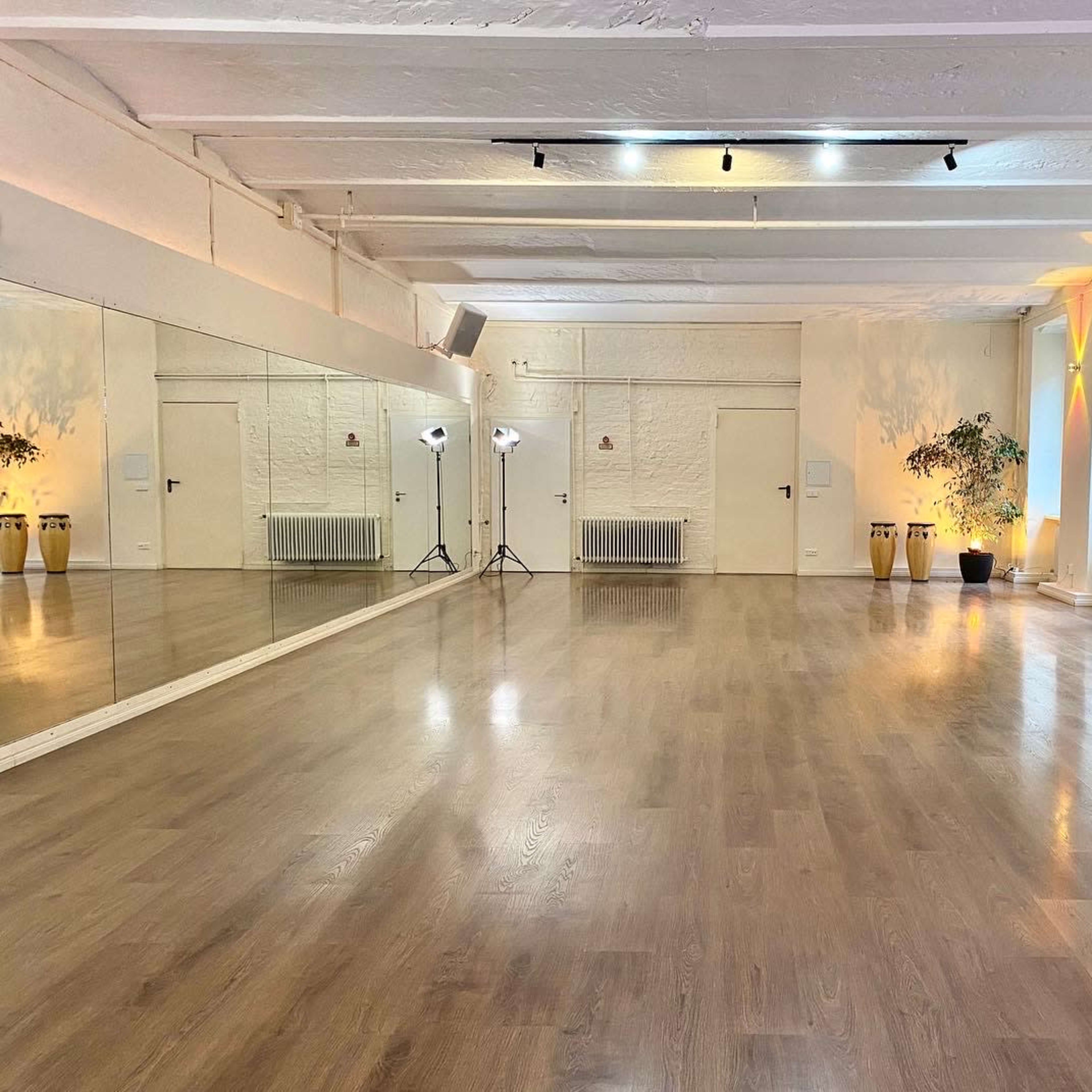 A spacious, well-lit dance studio features wooden flooring, mirrors on one wall, and soft lighting along the other walls.