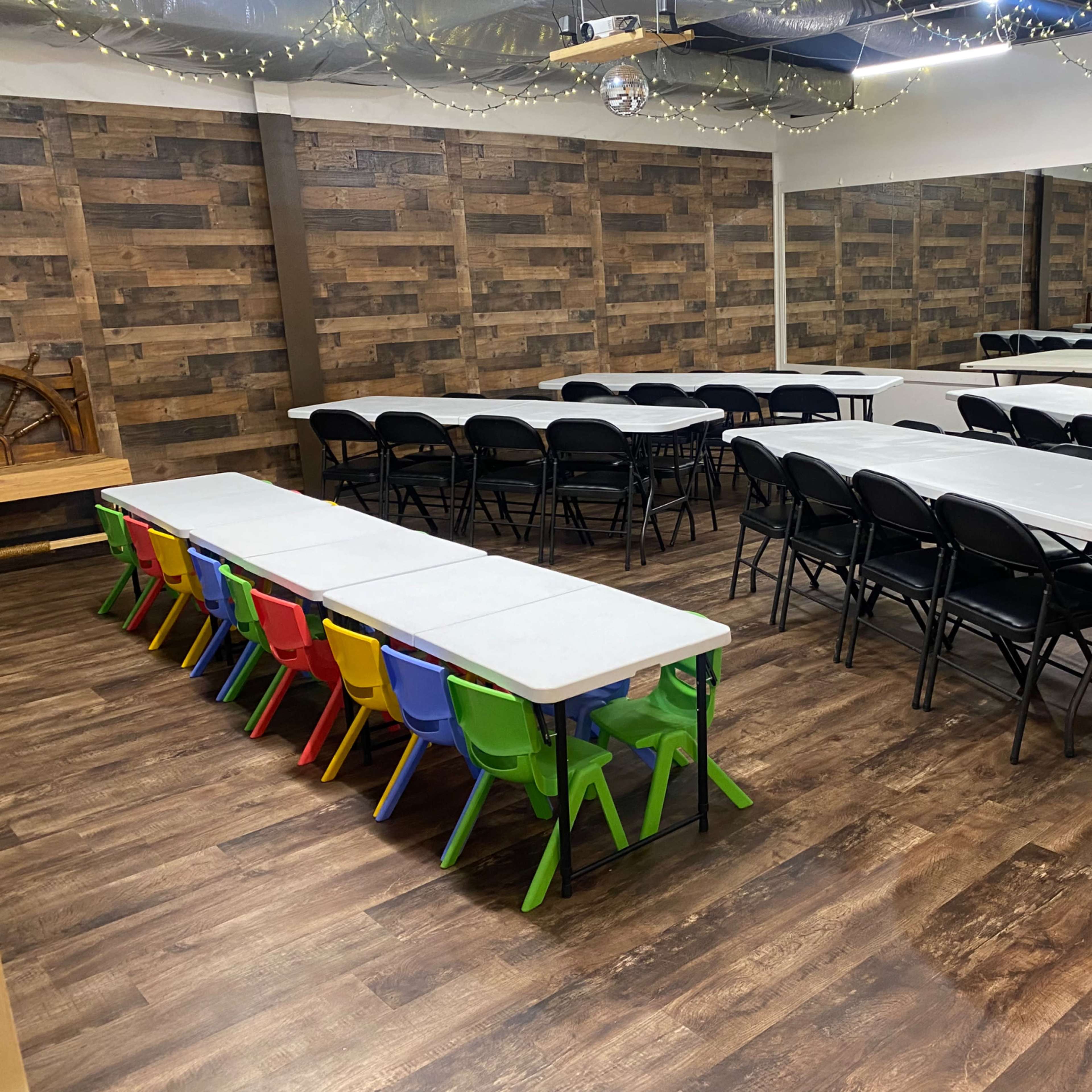 A spacious room features several rows of folding tables and chairs, with colorful plastic chairs lined up at one table against a wooden wall.