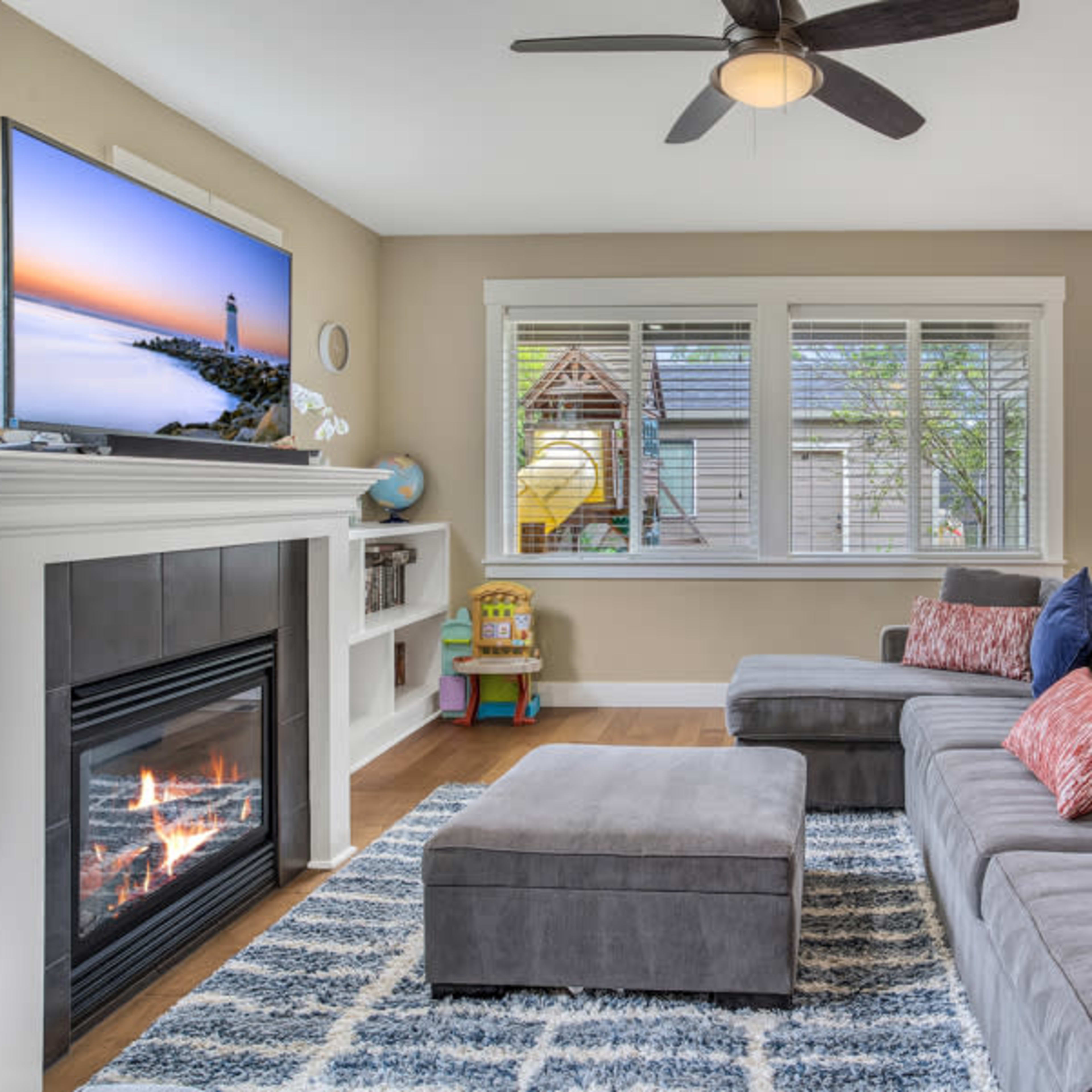 The living room features a gray sectional sofa, a television mounted above a fireplace, and a dining area with modern chairs and a visible play structure outside.