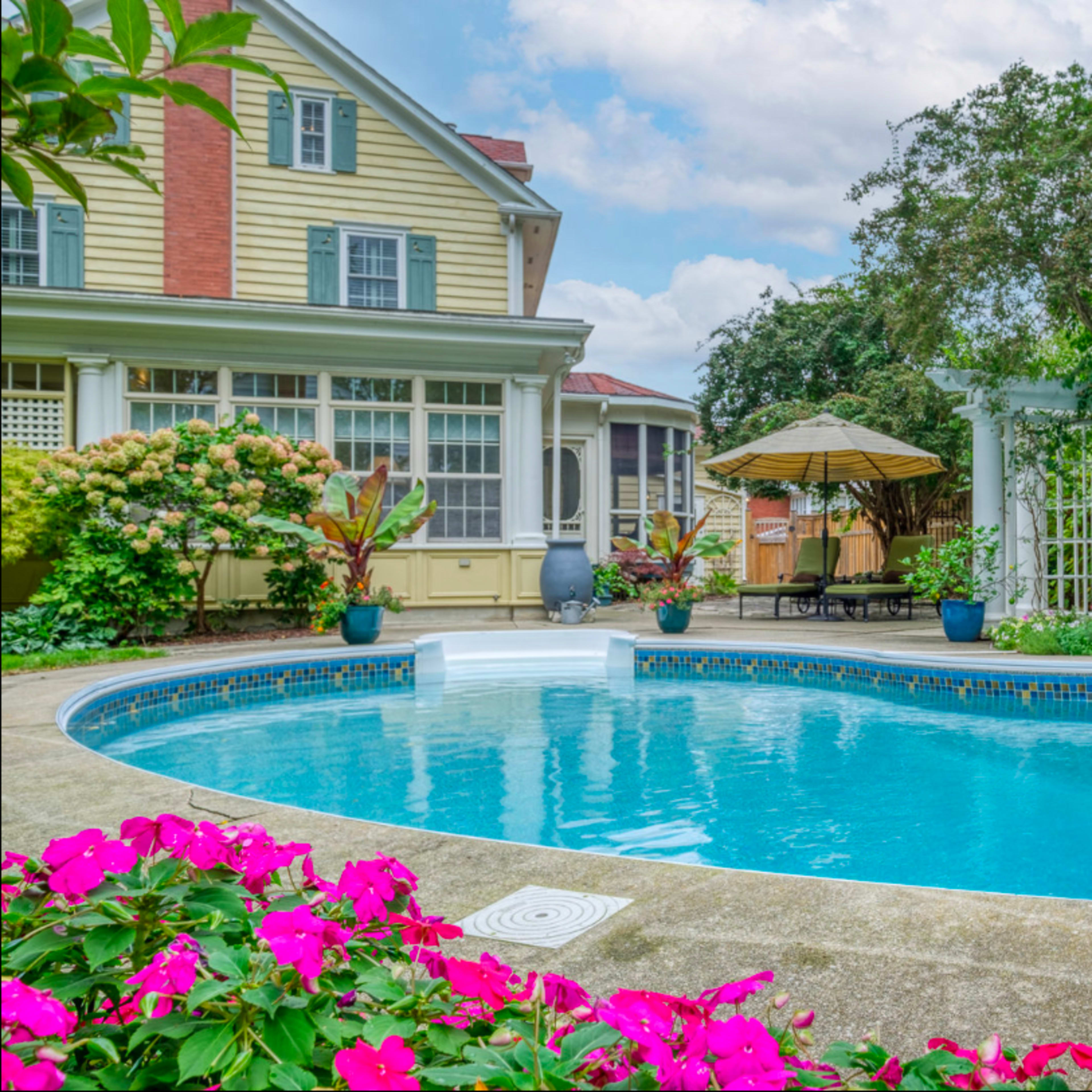 A clear swimming pool surrounded by colorful flowers and lush greenery, with a house and patio area in the background.