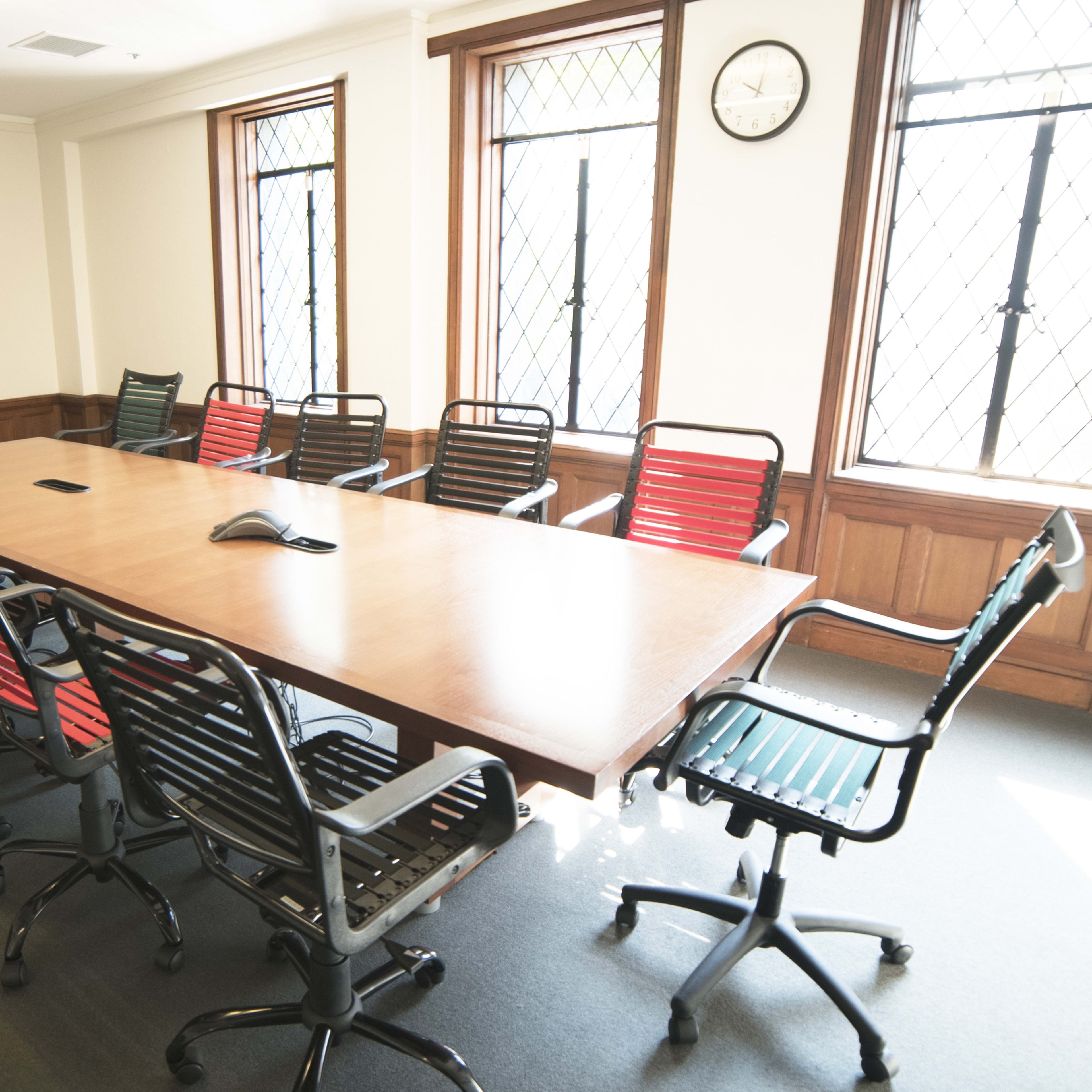 A long conference table surrounded by thirteen chairs is positioned in a meeting room with large windows and a wall-mounted television.