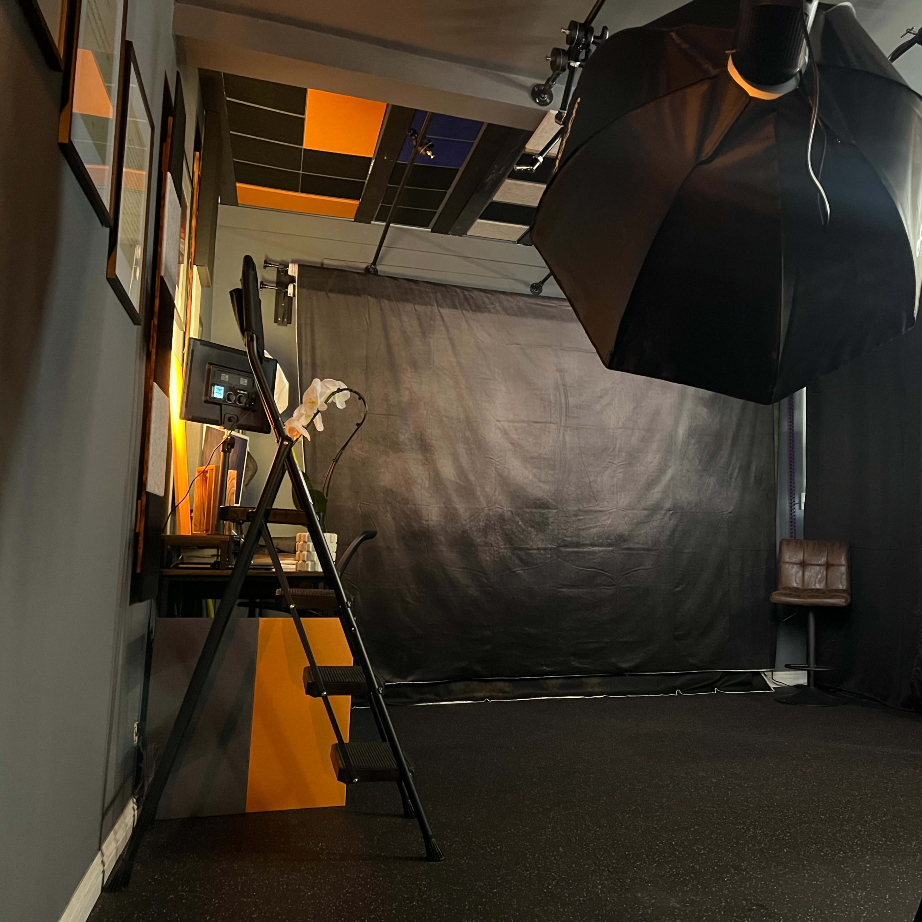 An indoor studio setup with a black backdrop, lighting equipment, a ladder, and a chair in the corner.