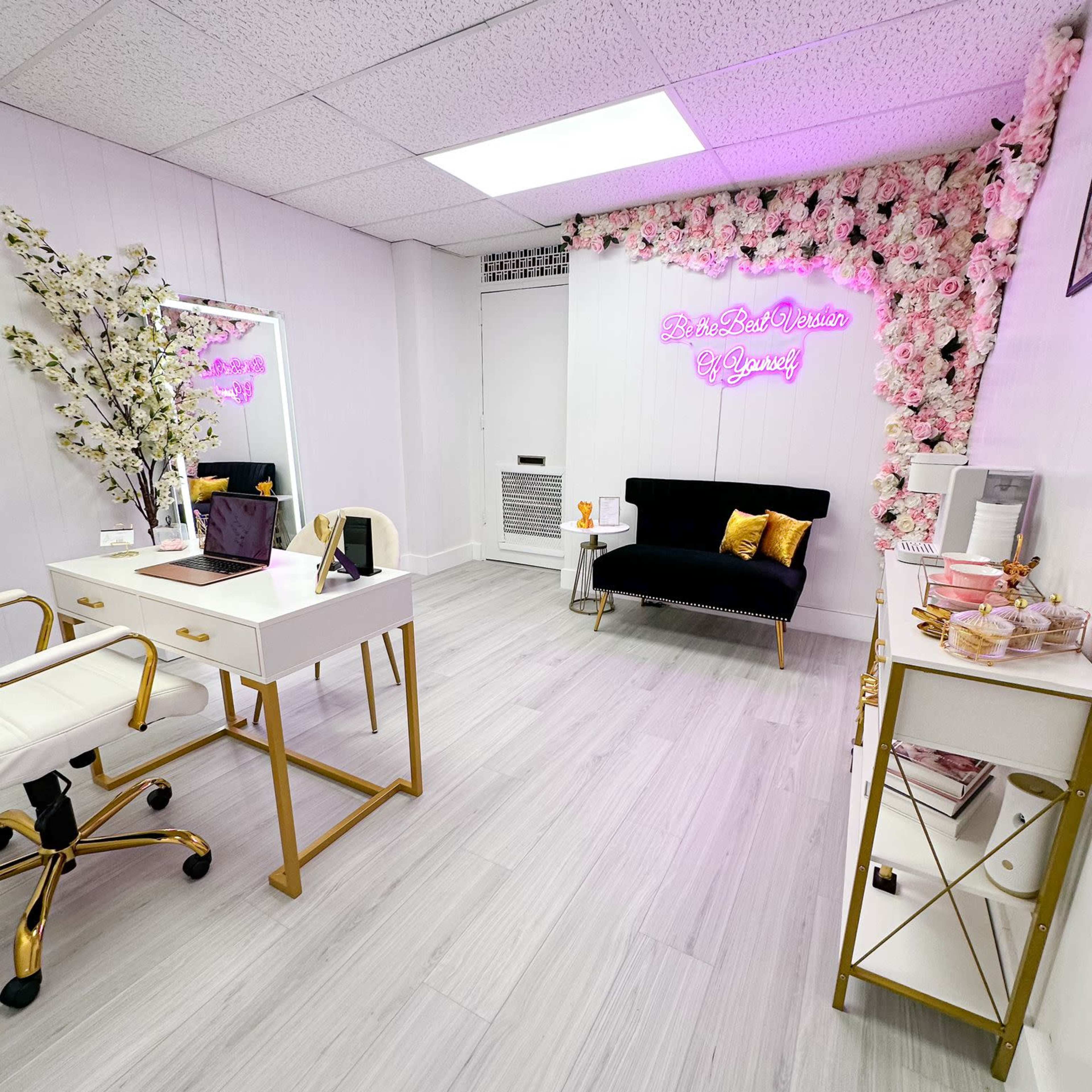 A modern beauty studio with a white and gold aesthetic, featuring a black sofa, decorative floral wall, and neon signage.