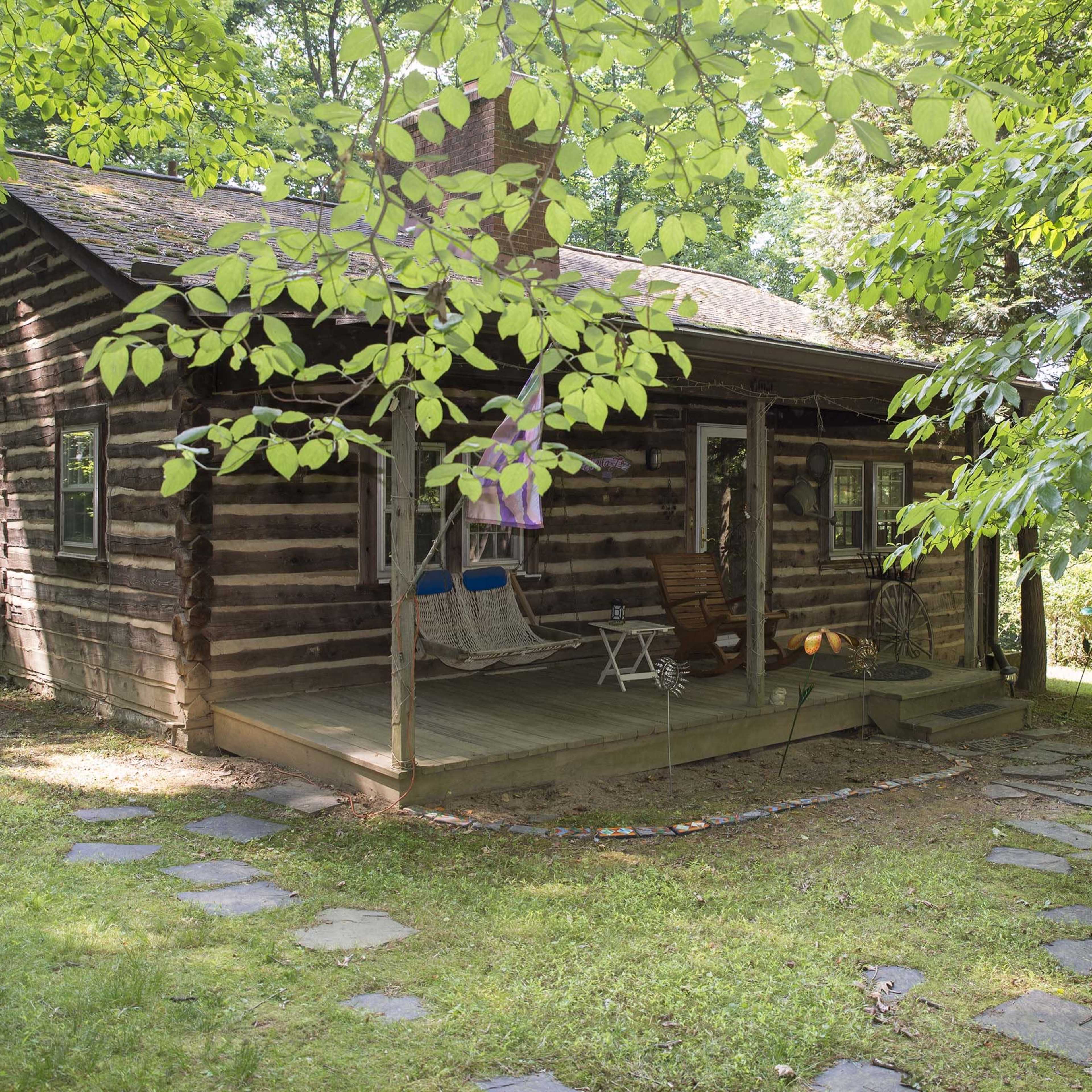 A rustic log cabin is surrounded by lush greenery and features a porch with seating and a stone walkway.