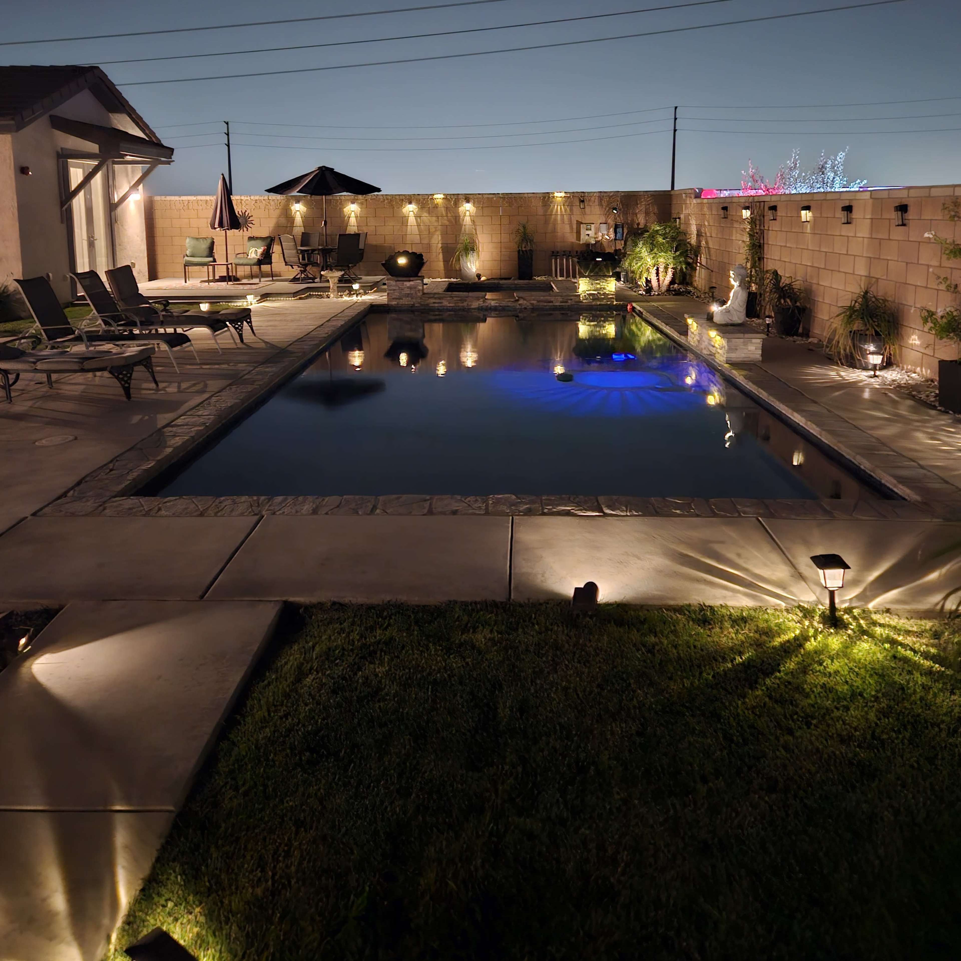 A well-lit backyard features a swimming pool surrounded by loungers and a patio umbrella, set against a clear night sky.