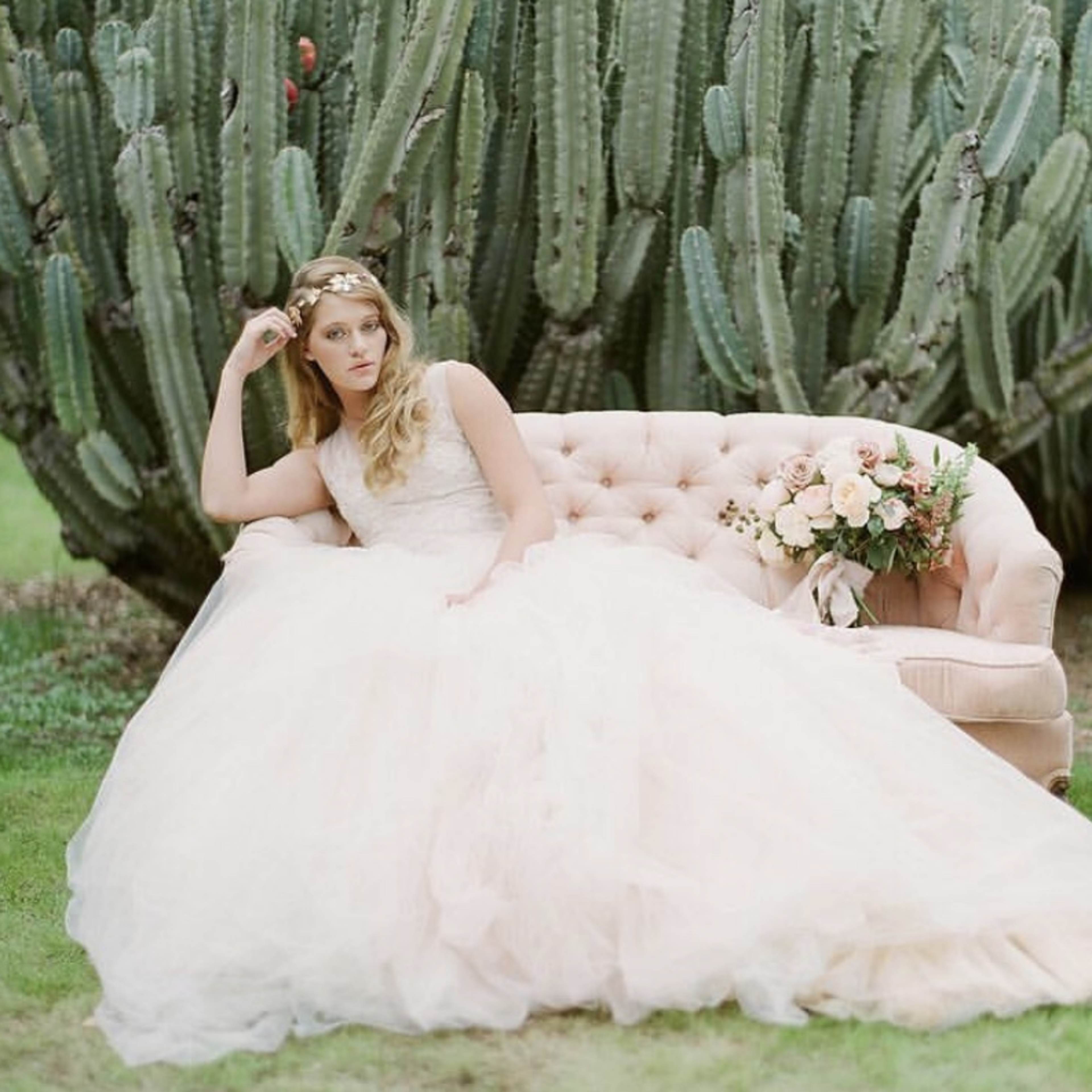 A bride in a flowing gown sits on a light pink couch beside a large cactus in a green outdoor setting.