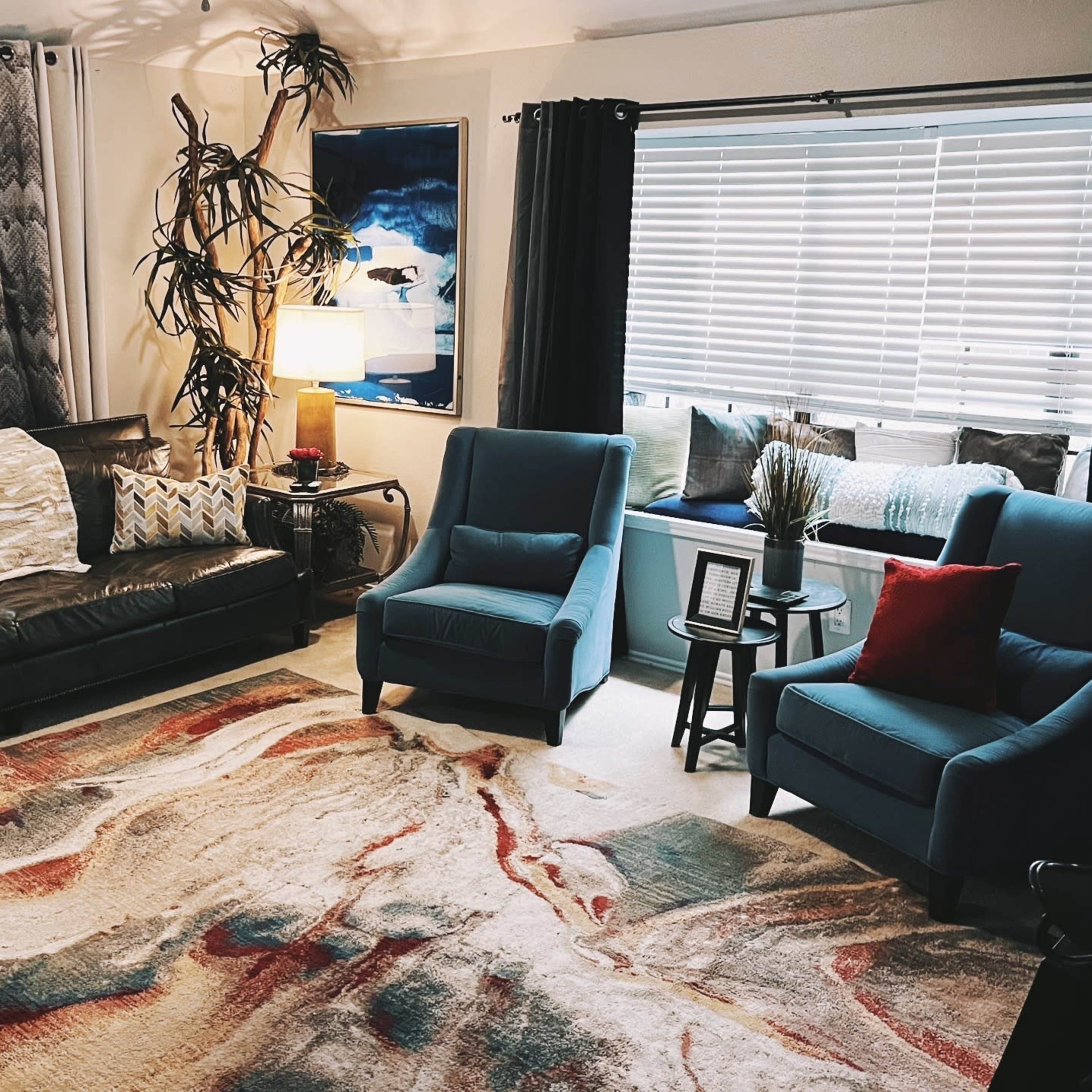 A living room with a black leather sofa, two blue chairs, a large area rug, and a window seat with cushions.