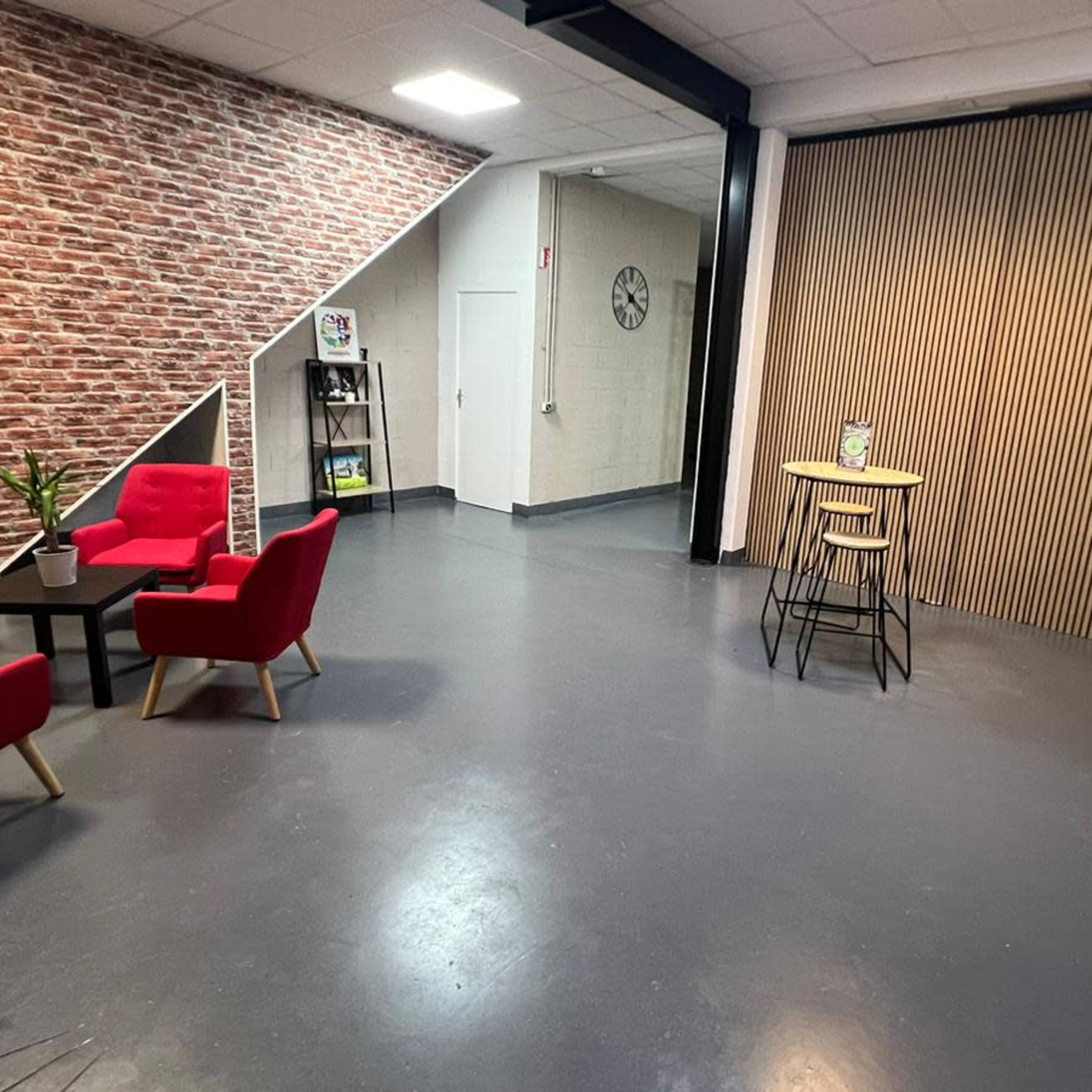 A spacious room with a brick wall, featuring red armchairs grouped around a black table and a wooden counter with bar stools against a backdrop of wooden slats.