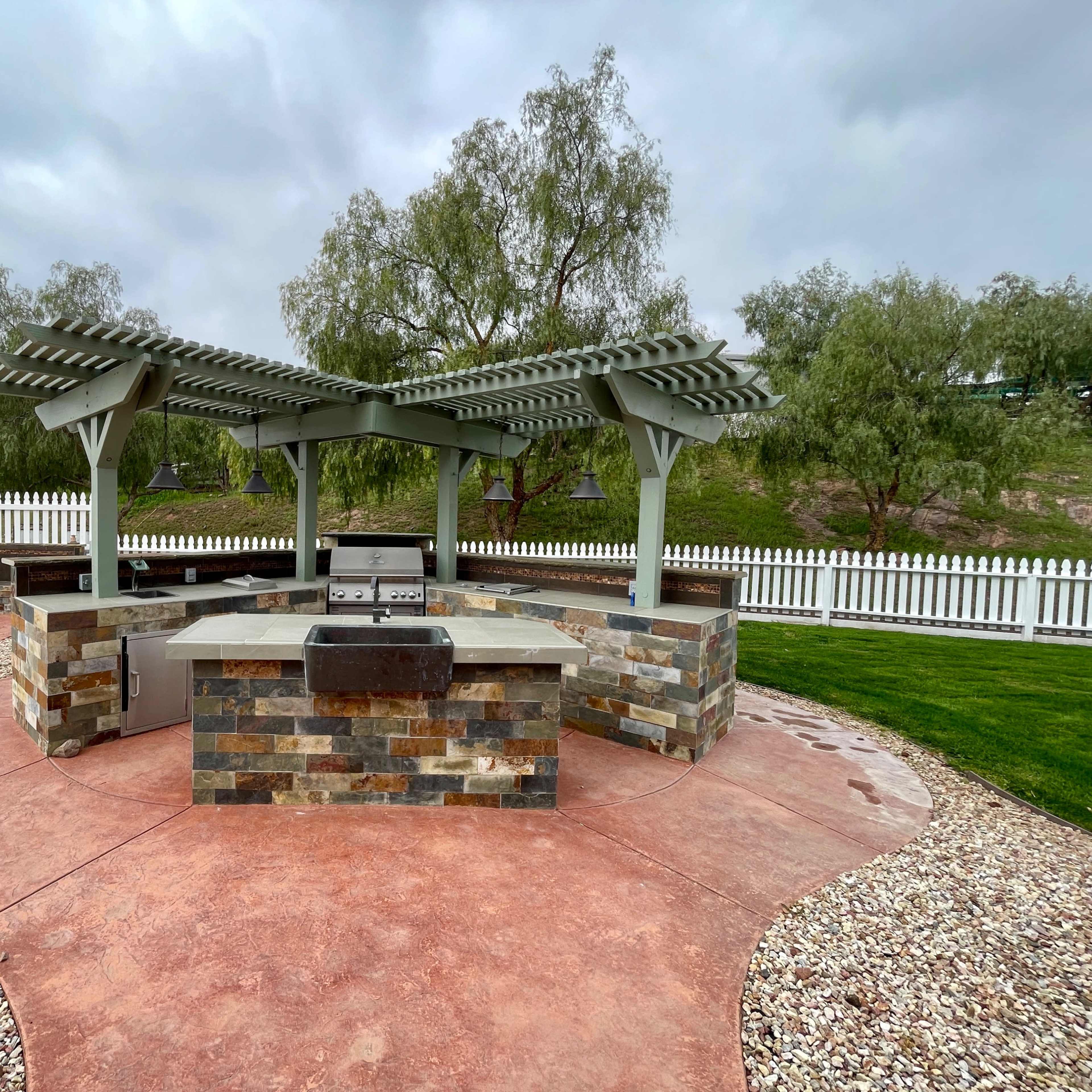An outdoor kitchen area featuring a stone-built barbecue station under a pergola, surrounded by a landscaped yard with a white picket fence.