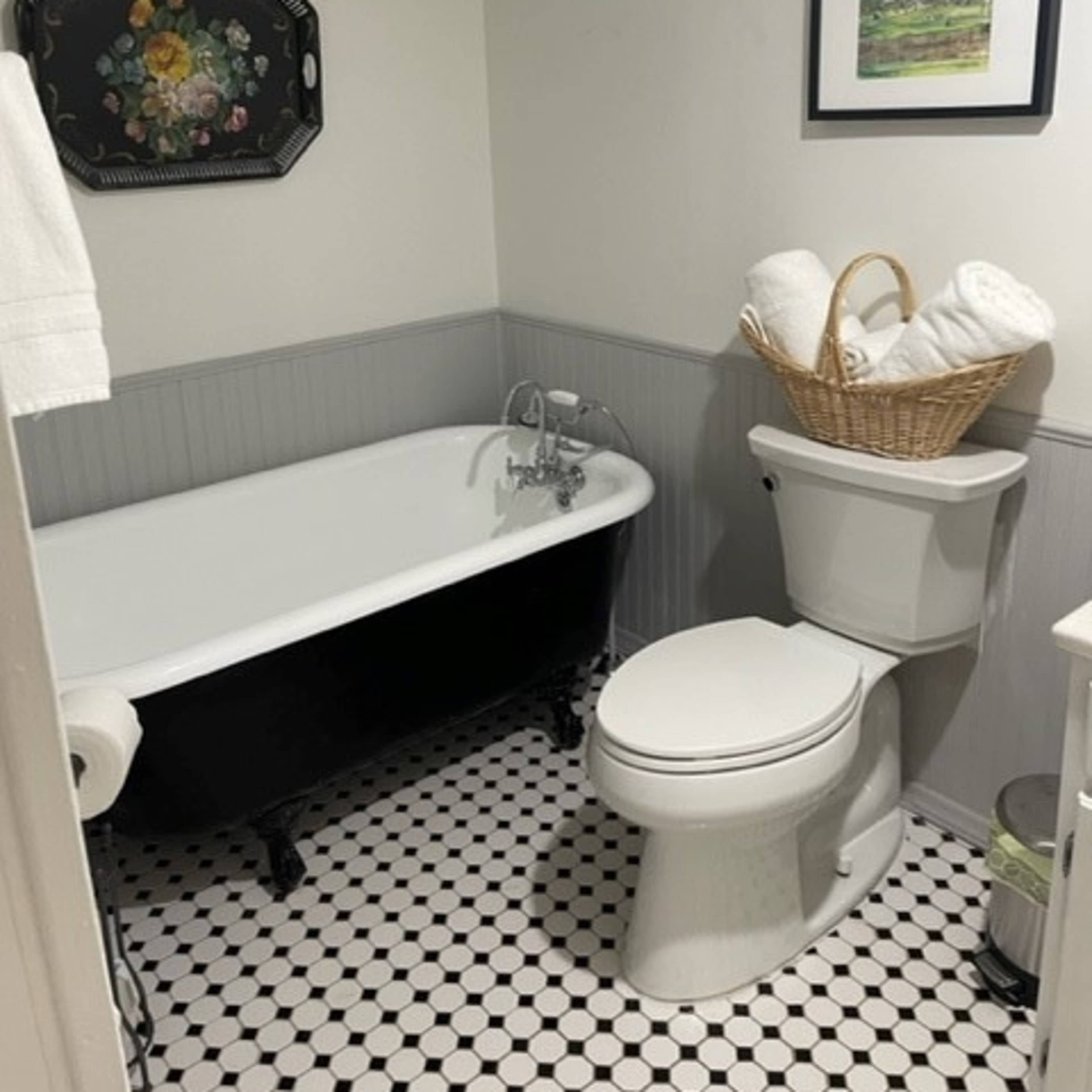 A bathroom featuring a black-clawed bathtub, a white toilet, a wicker basket filled with towels, and a small framed picture on the wall.