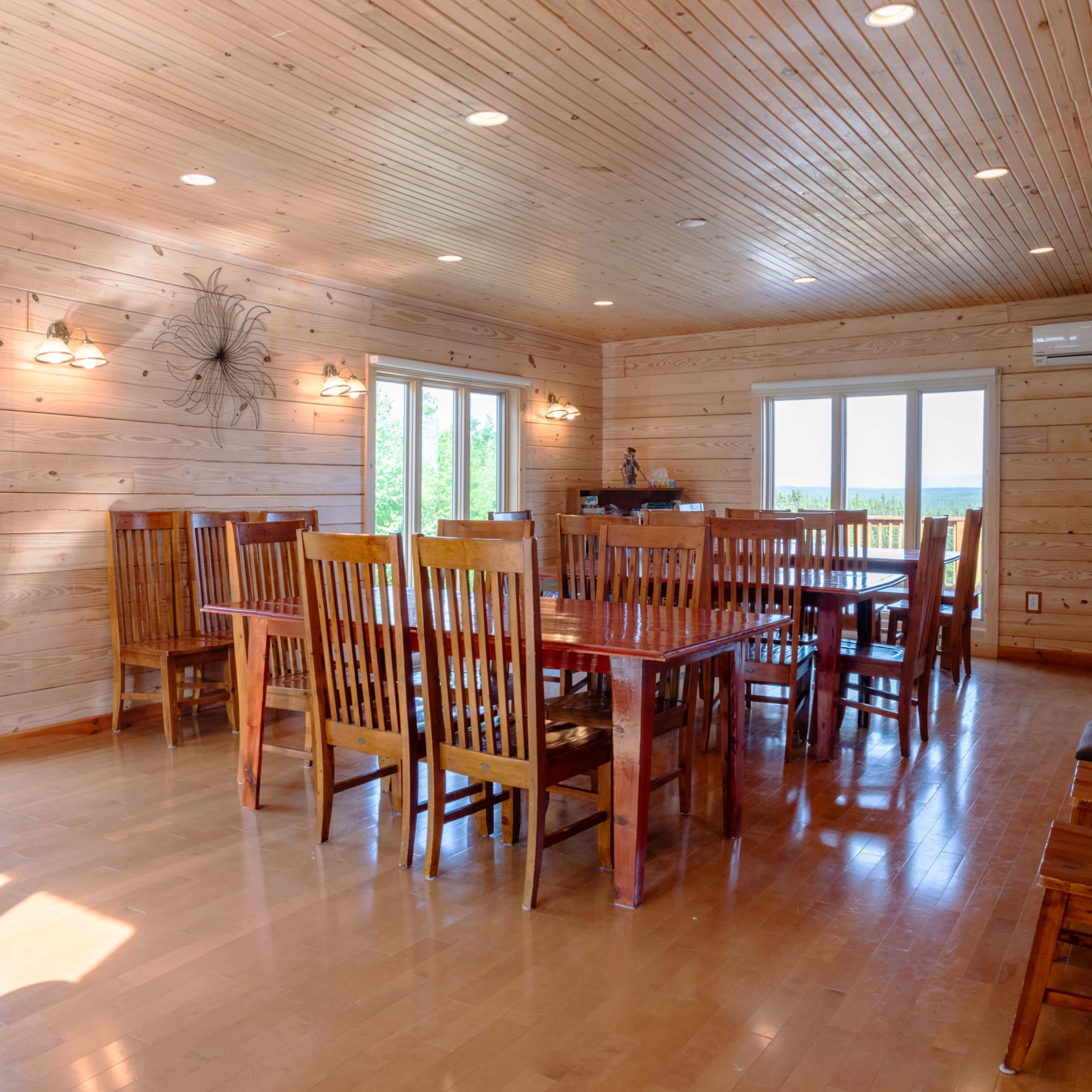 A spacious wooden dining area featuring multiple large tables surrounded by wooden chairs, with windows allowing natural light to illuminate the room.