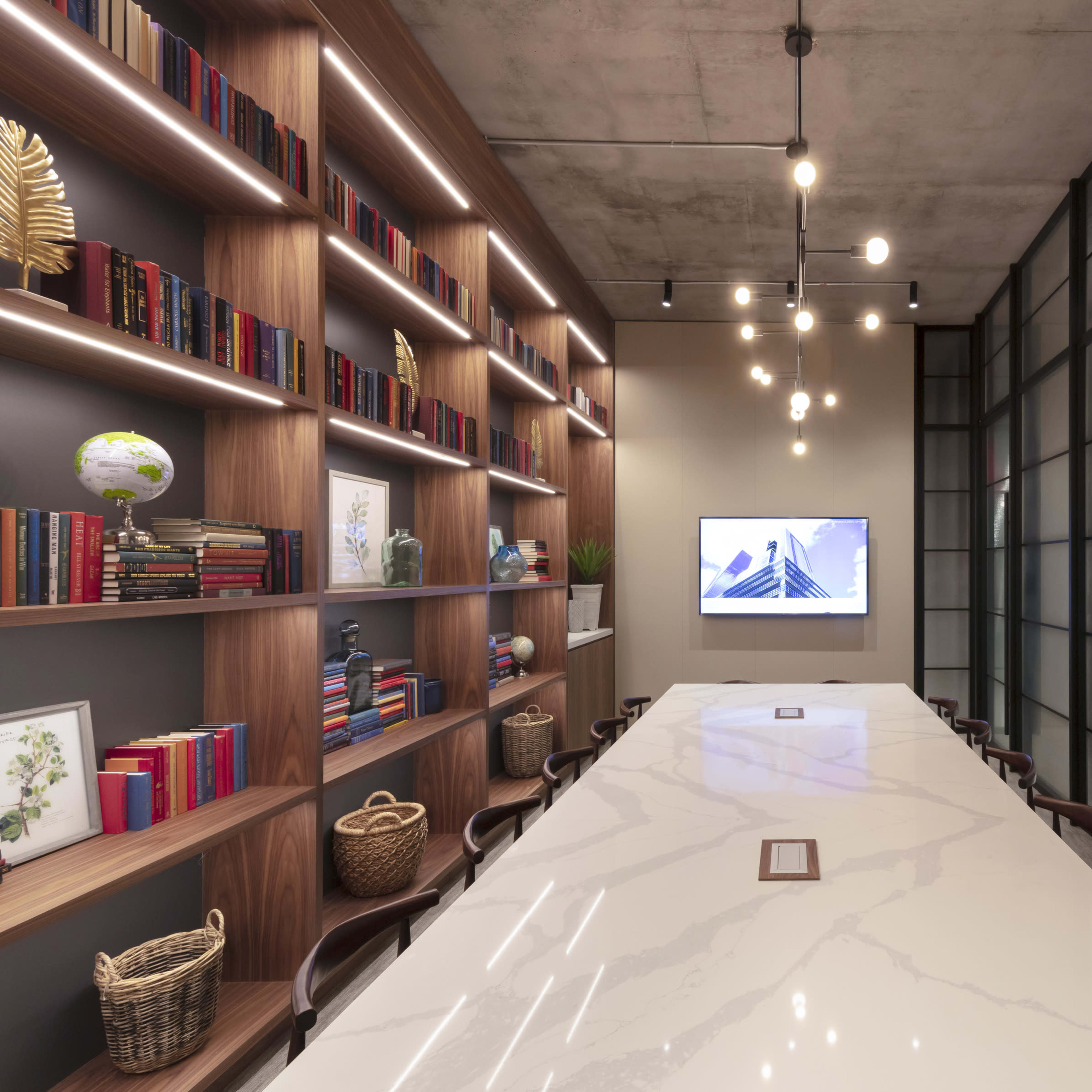 A minimalist conference room features a long marble table, wall-mounted screens, and shelves lined with books and decorative items.