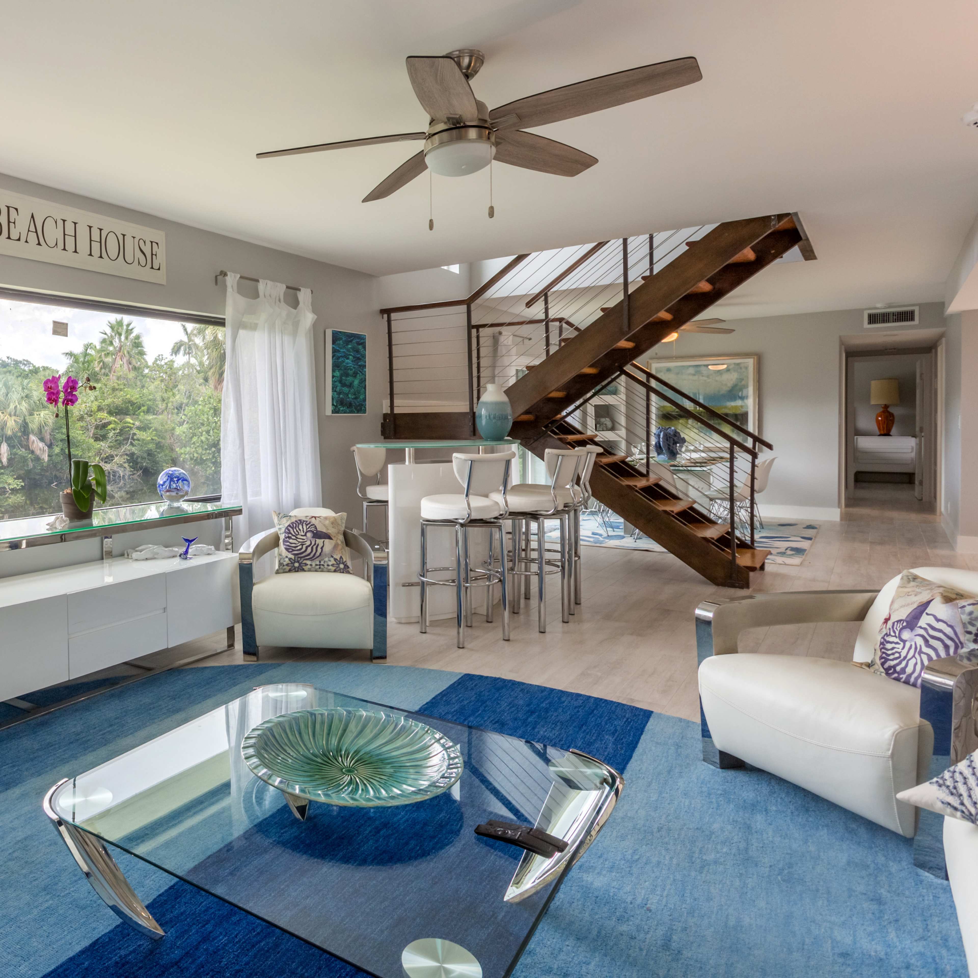 A modern living room with a large window, a blue area rug, a white coffee table, and a staircase leading to an upper level.
