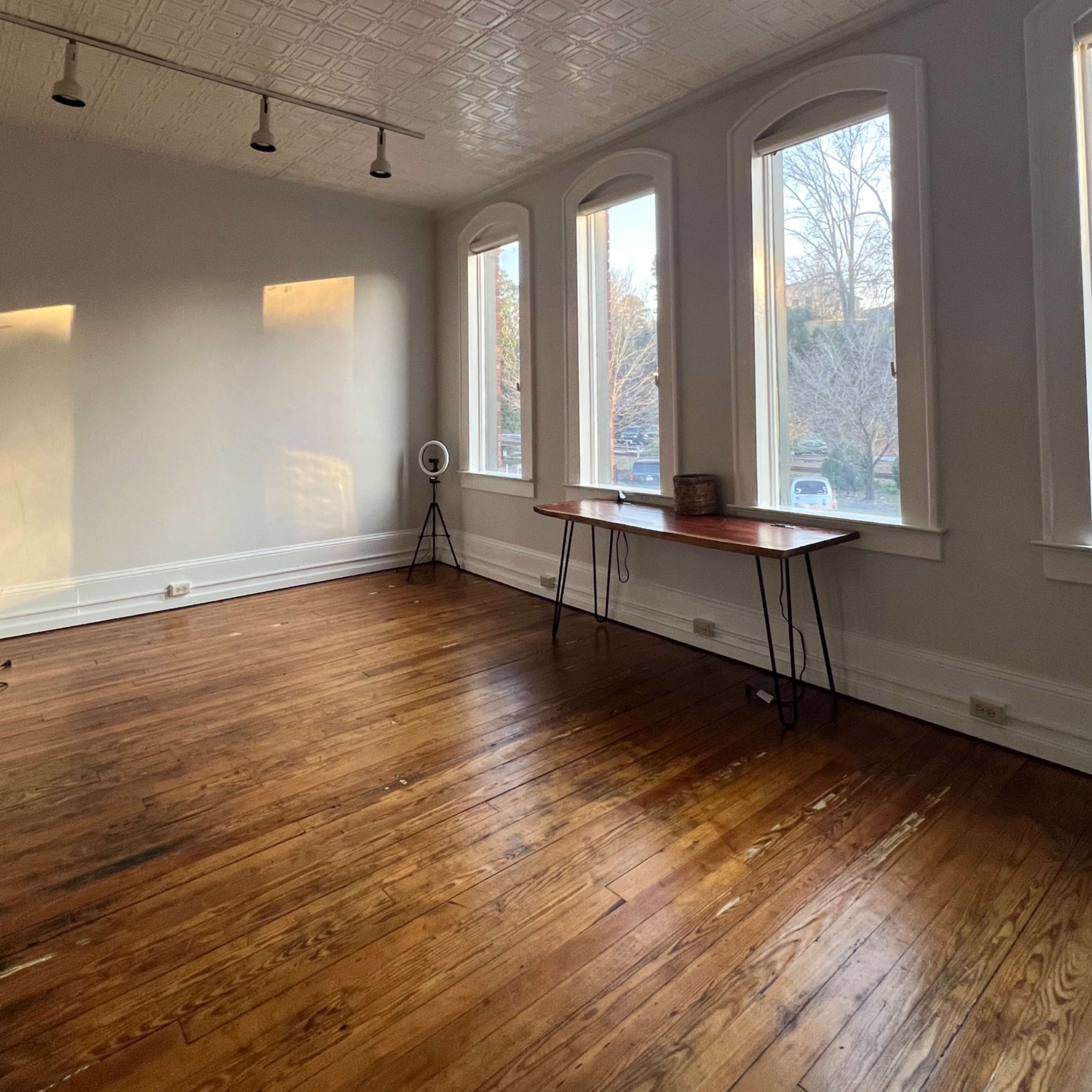 A spacious, empty room with wooden floors, a table against the wall, and large windows letting in natural light.