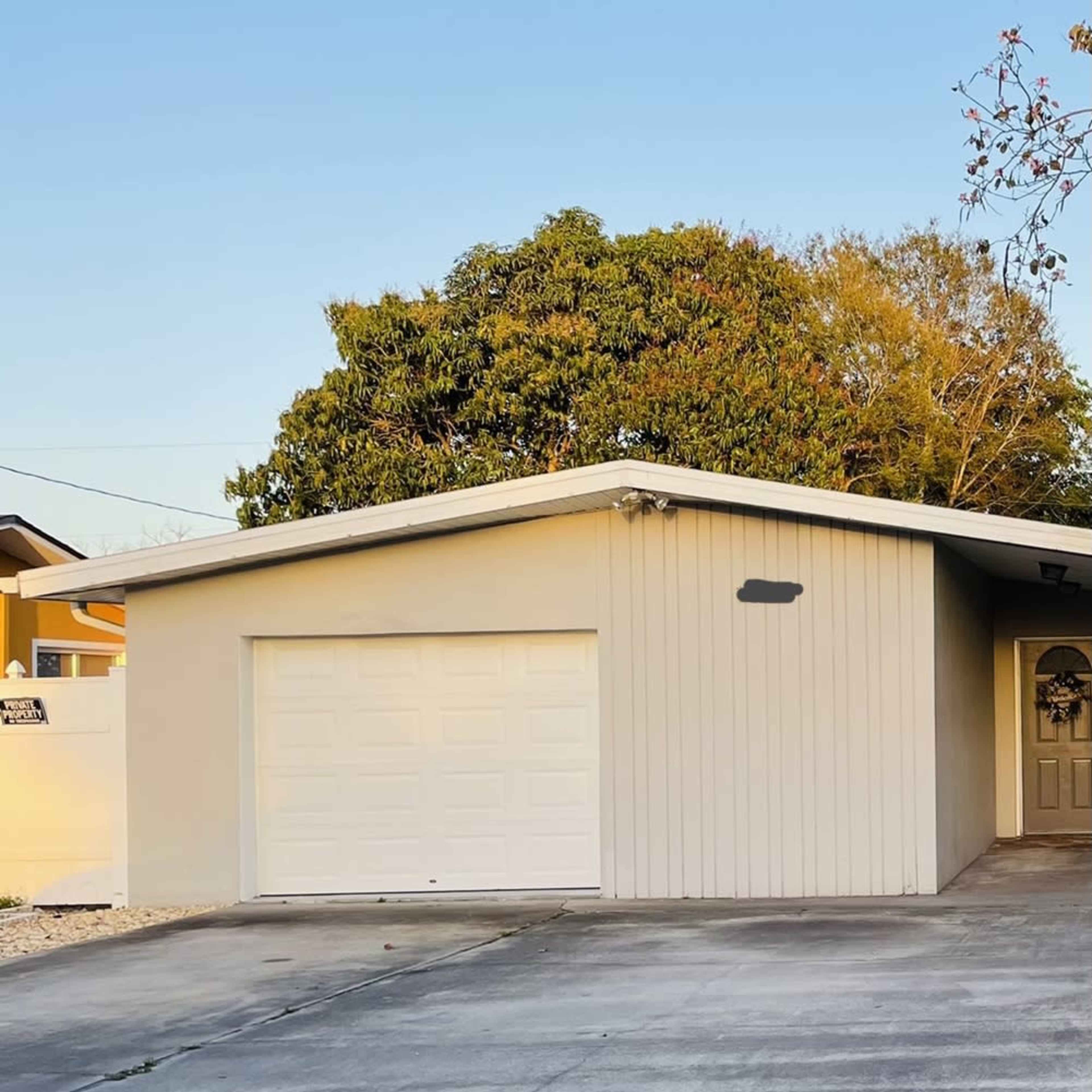 A single-story house with a gray exterior and a white garage door is located on a concrete driveway under a clear blue sky.