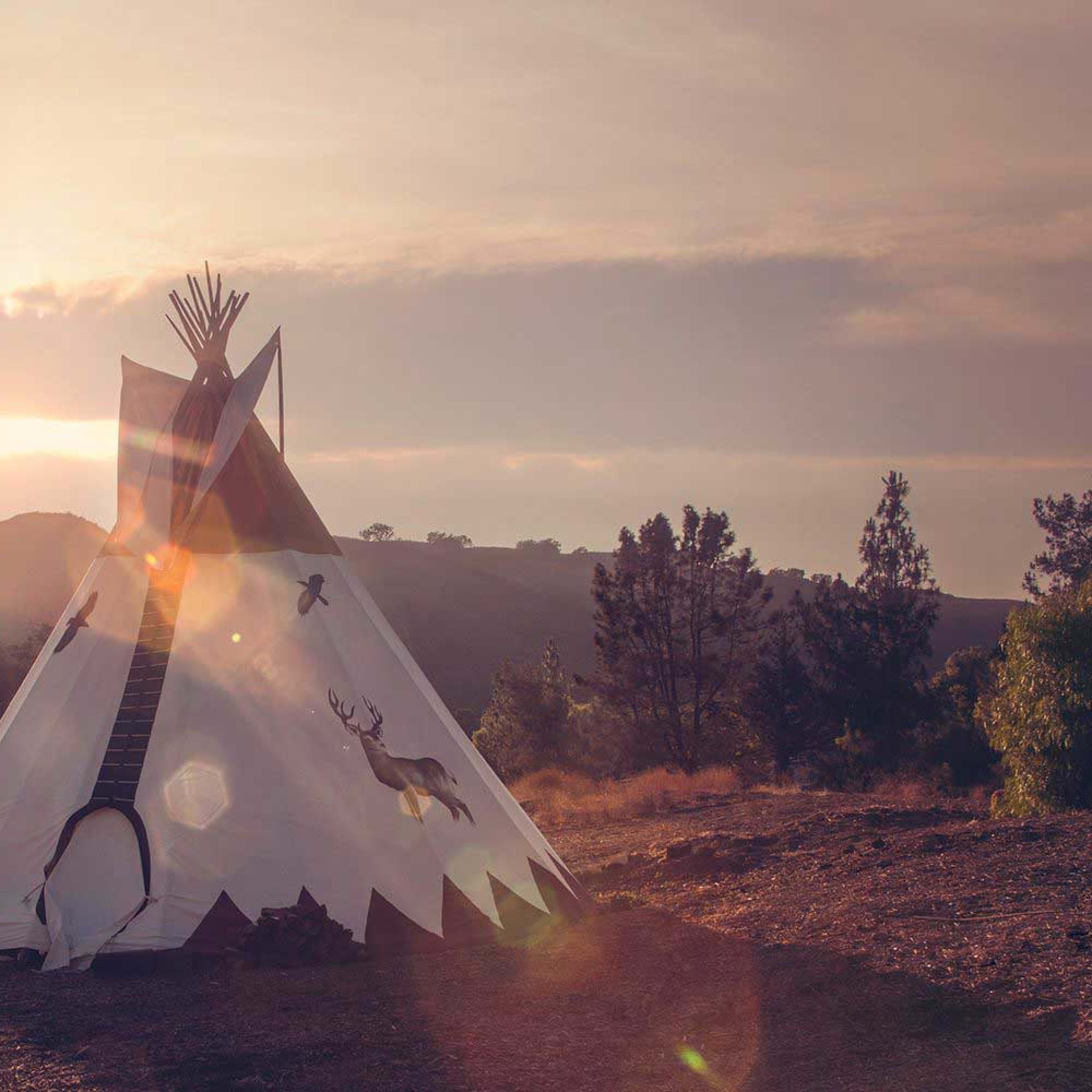 A teepee is set against a backdrop of mountains under a sunset sky.