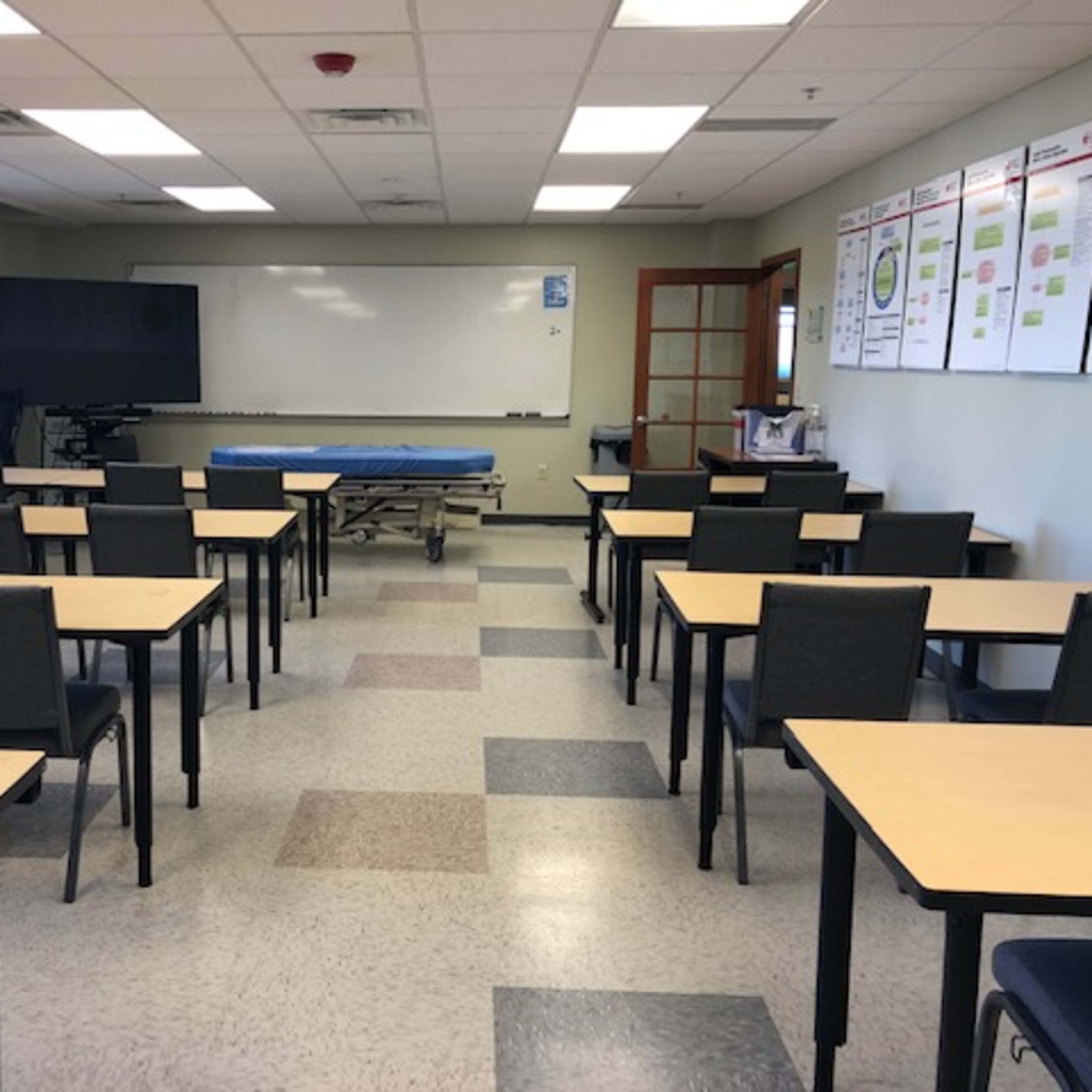 A classroom with rows of tables and chairs, a whiteboard at the front, and a mobile cart positioned against the wall.