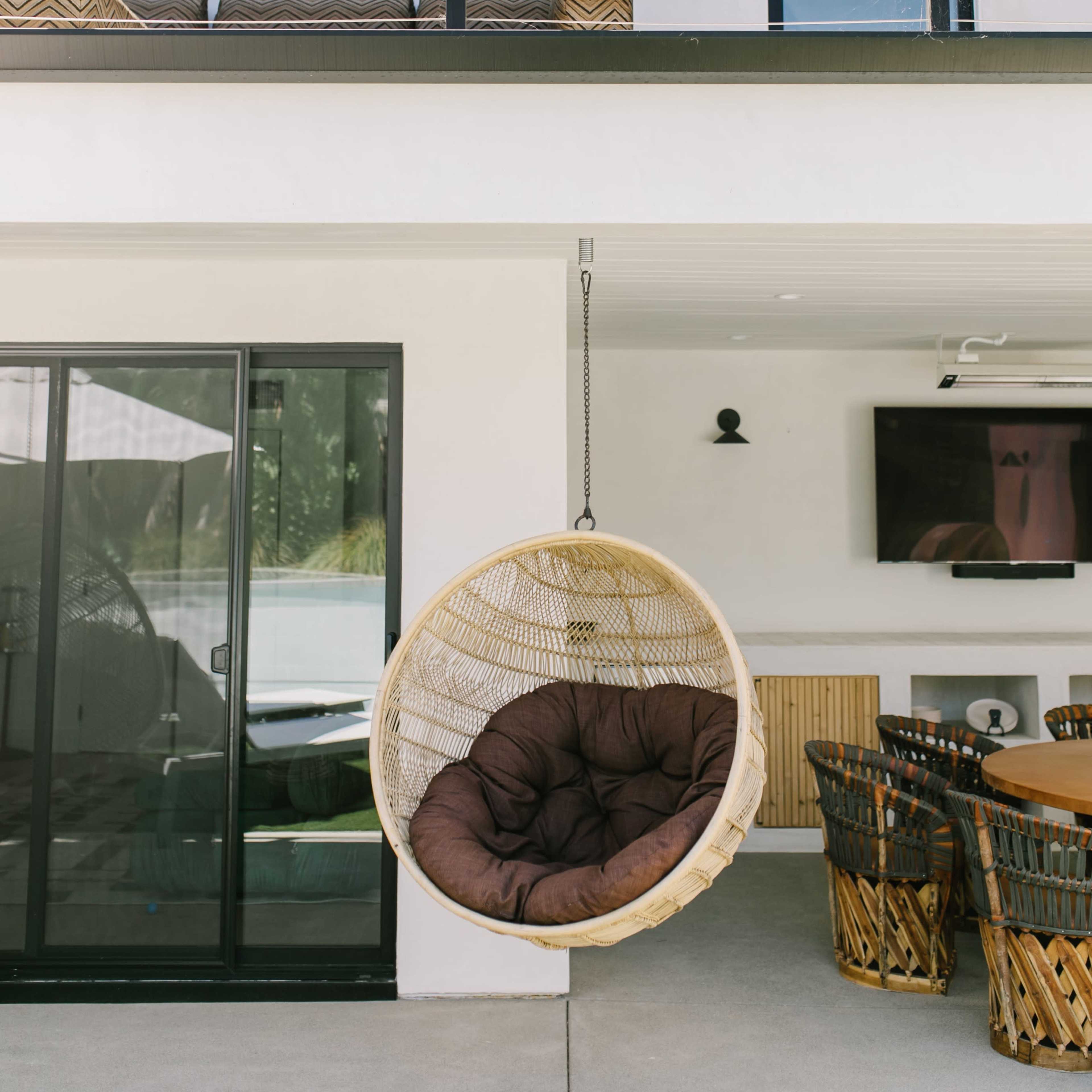 A hanging chair is suspended near a modern outdoor dining area, with a two-story building featuring large windows in the background.