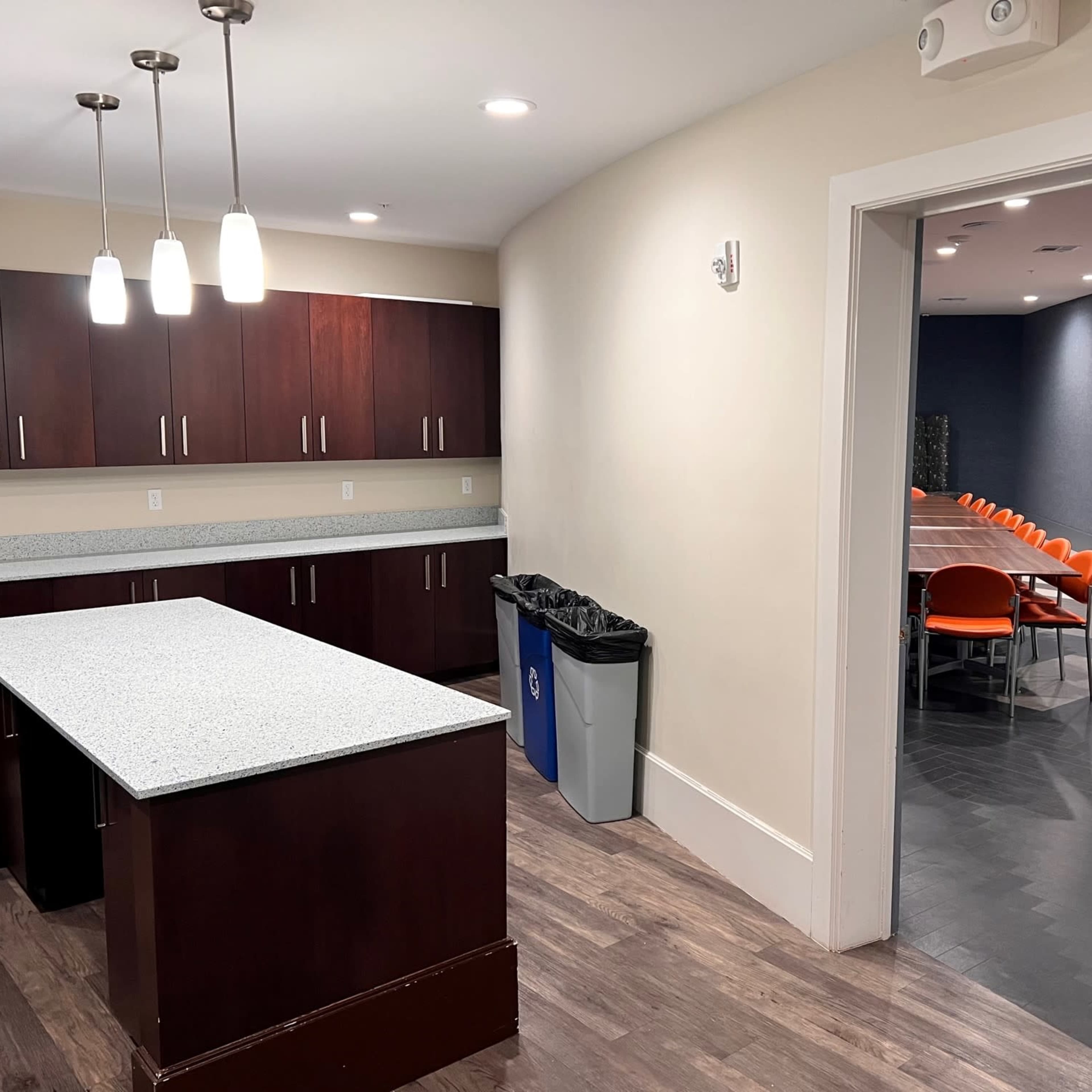 A modern kitchen area with a granite countertop island, dark cabinetry, and an open doorway leading to a meeting room with orange chairs.
