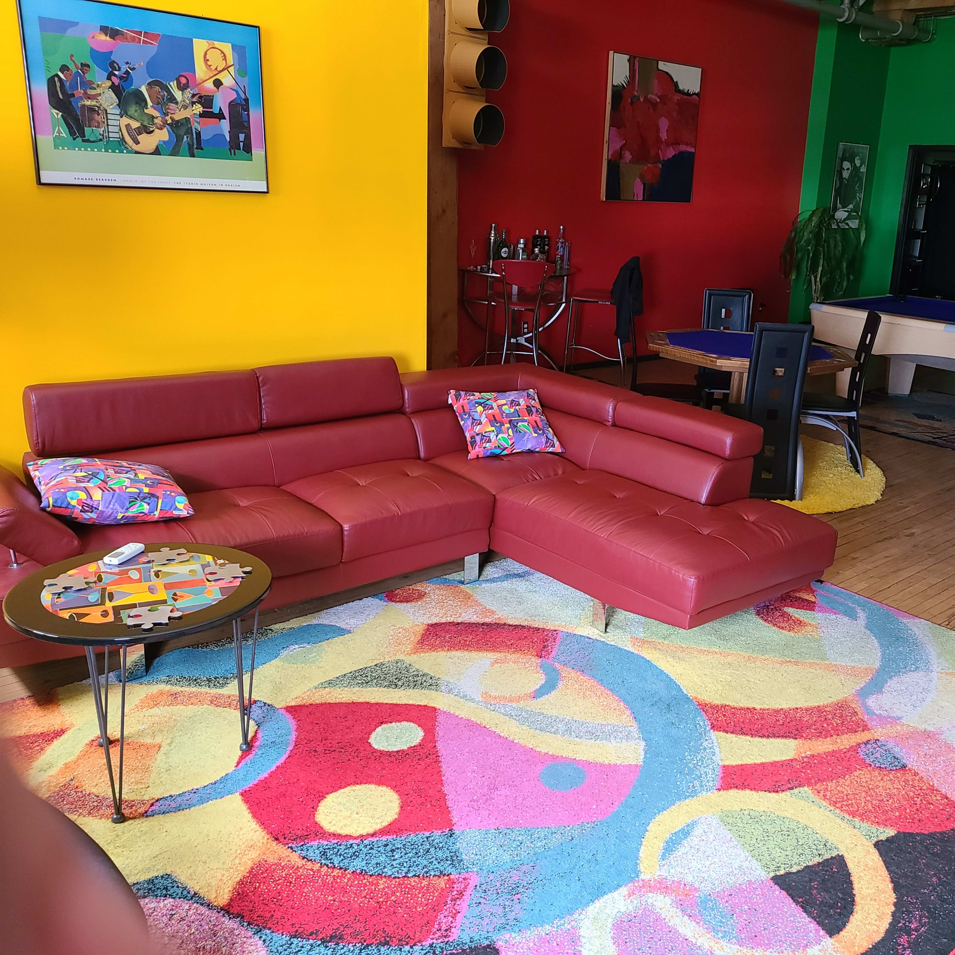 The scene features a vibrant living room with a red sectional sofa, a colorful area rug, and brightly painted yellow and green walls, along with a pool table and modern decor.