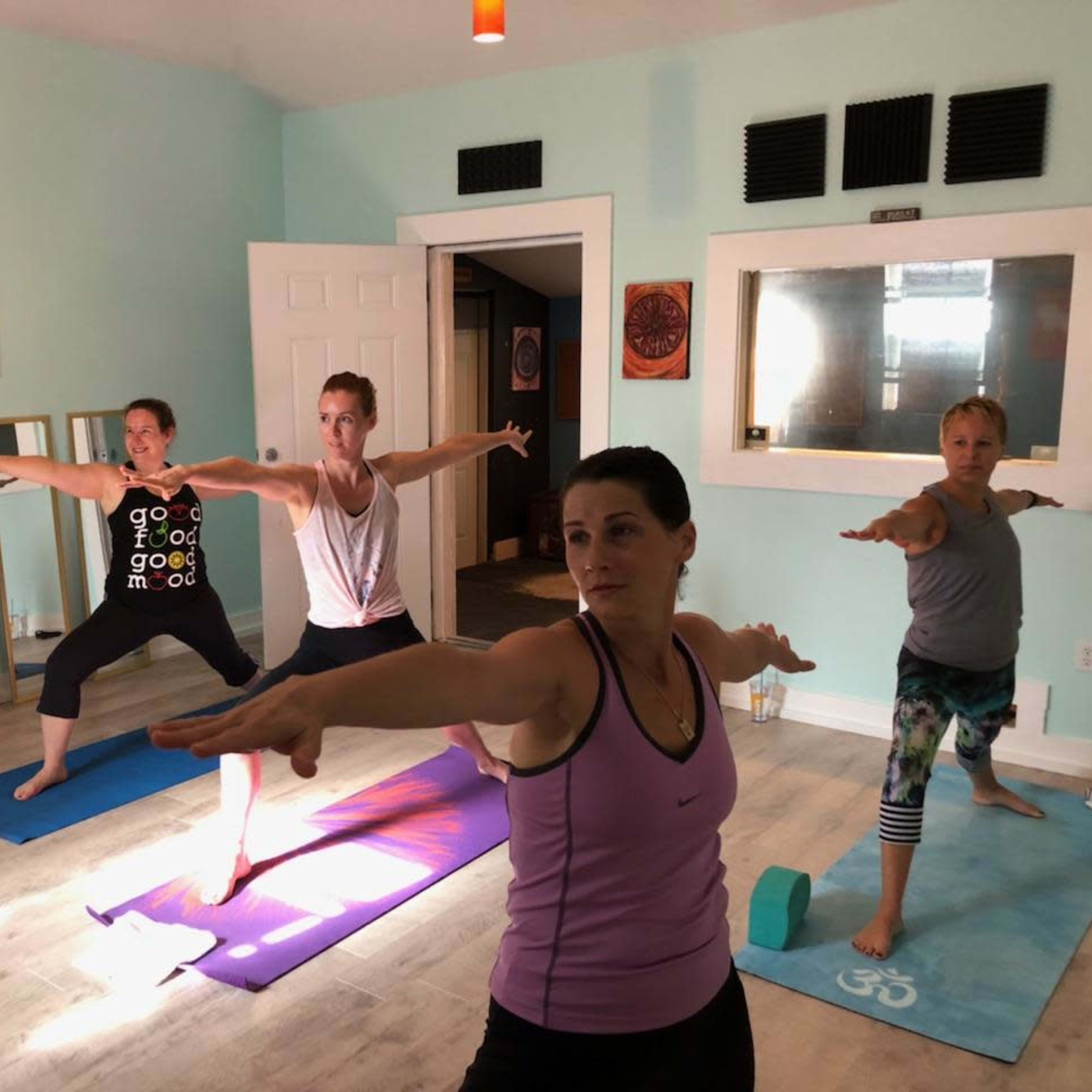 A group of four women practice yoga in a brightly lit studio with mirrors and colorful mats.