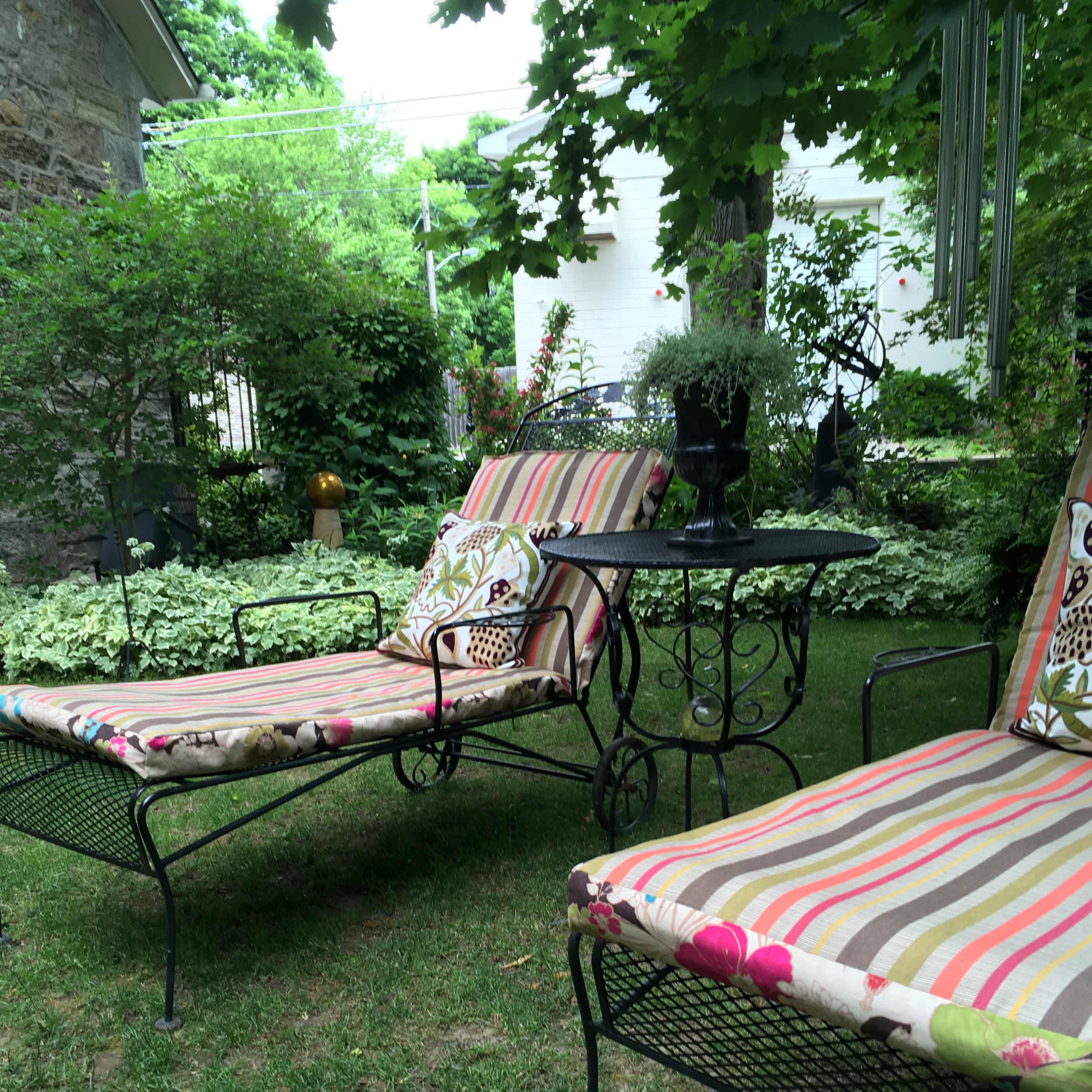 Two striped lounge chairs with decorative cushions sit beside a small metal table in a grassy garden surrounded by greenery.