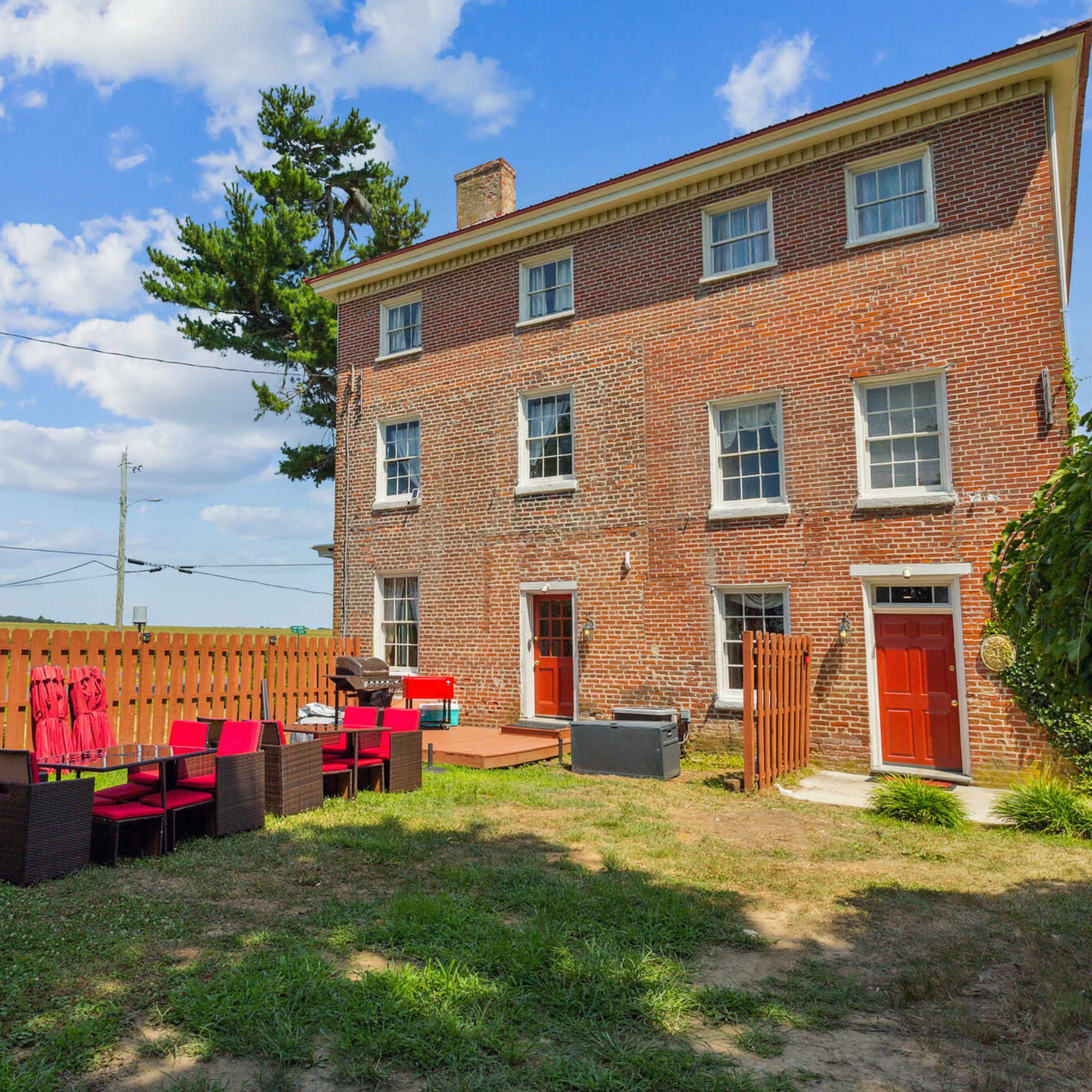 A three-story brick house with red doors and a fenced backyard features a seating area with red chairs and a hot tub.
