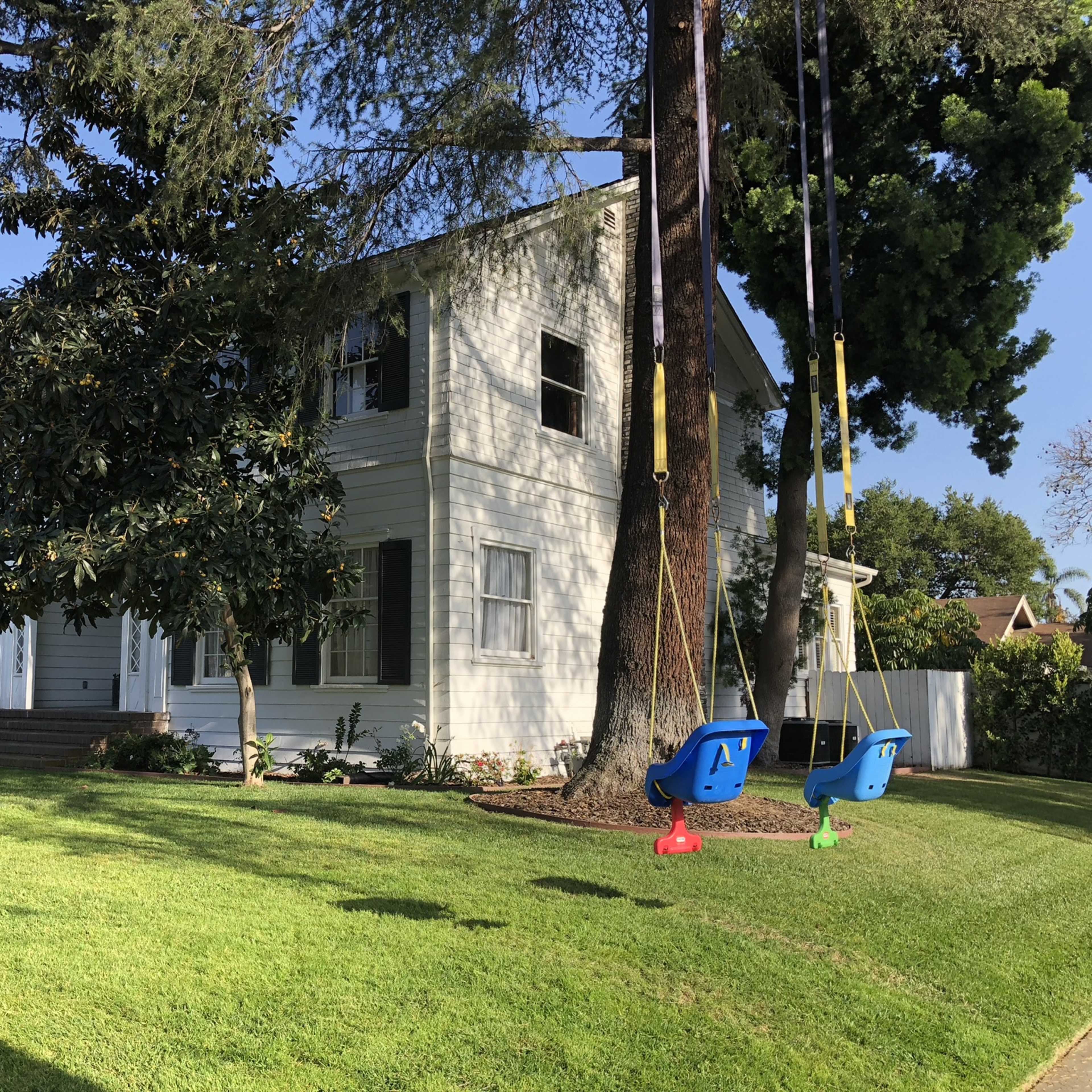 A two-story white house with green shutters sits on a well-maintained lawn, featuring two swings hanging from tree branches nearby.