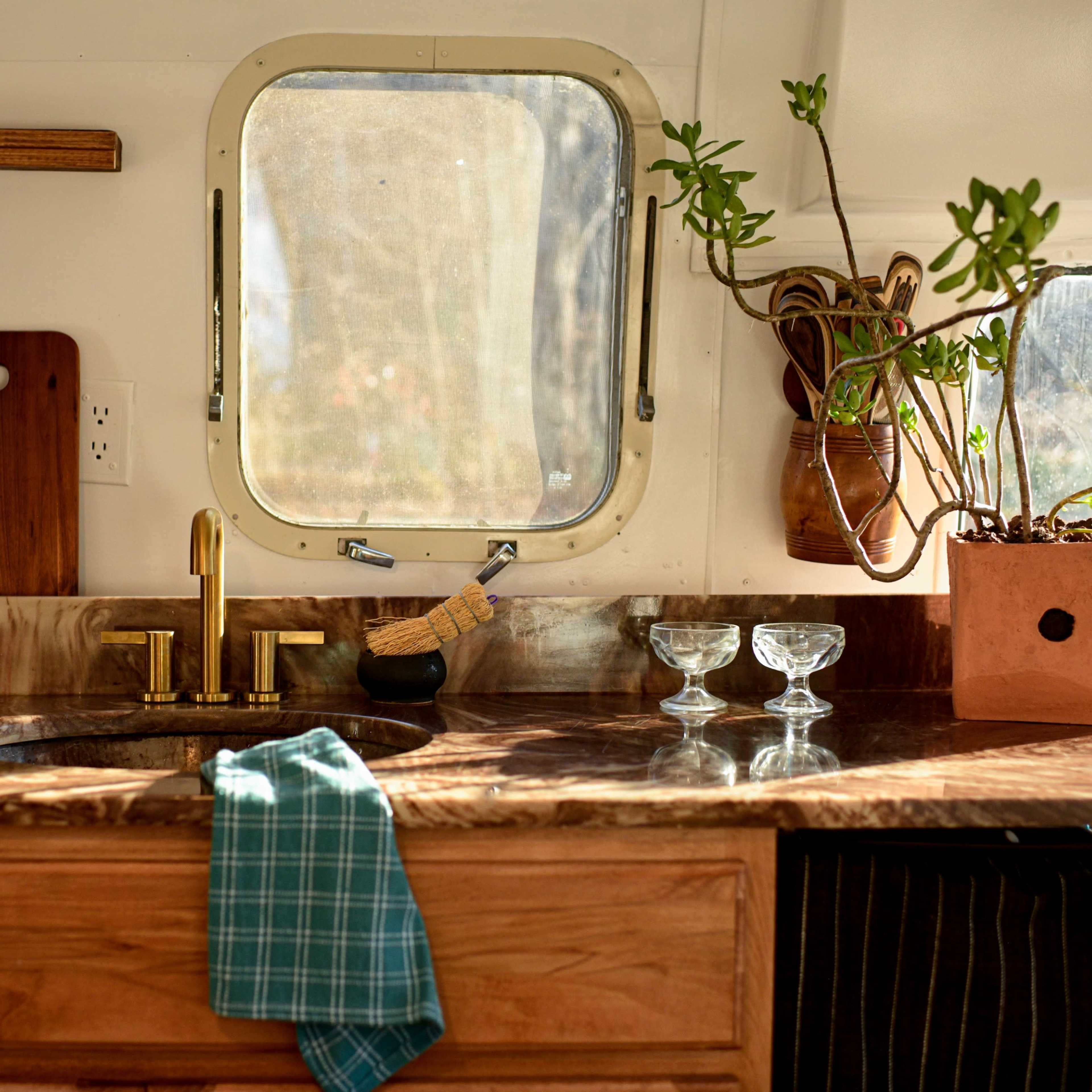 A kitchen countertop with a sink, gold faucet, wooden cutting board, and potted plant near a window.
