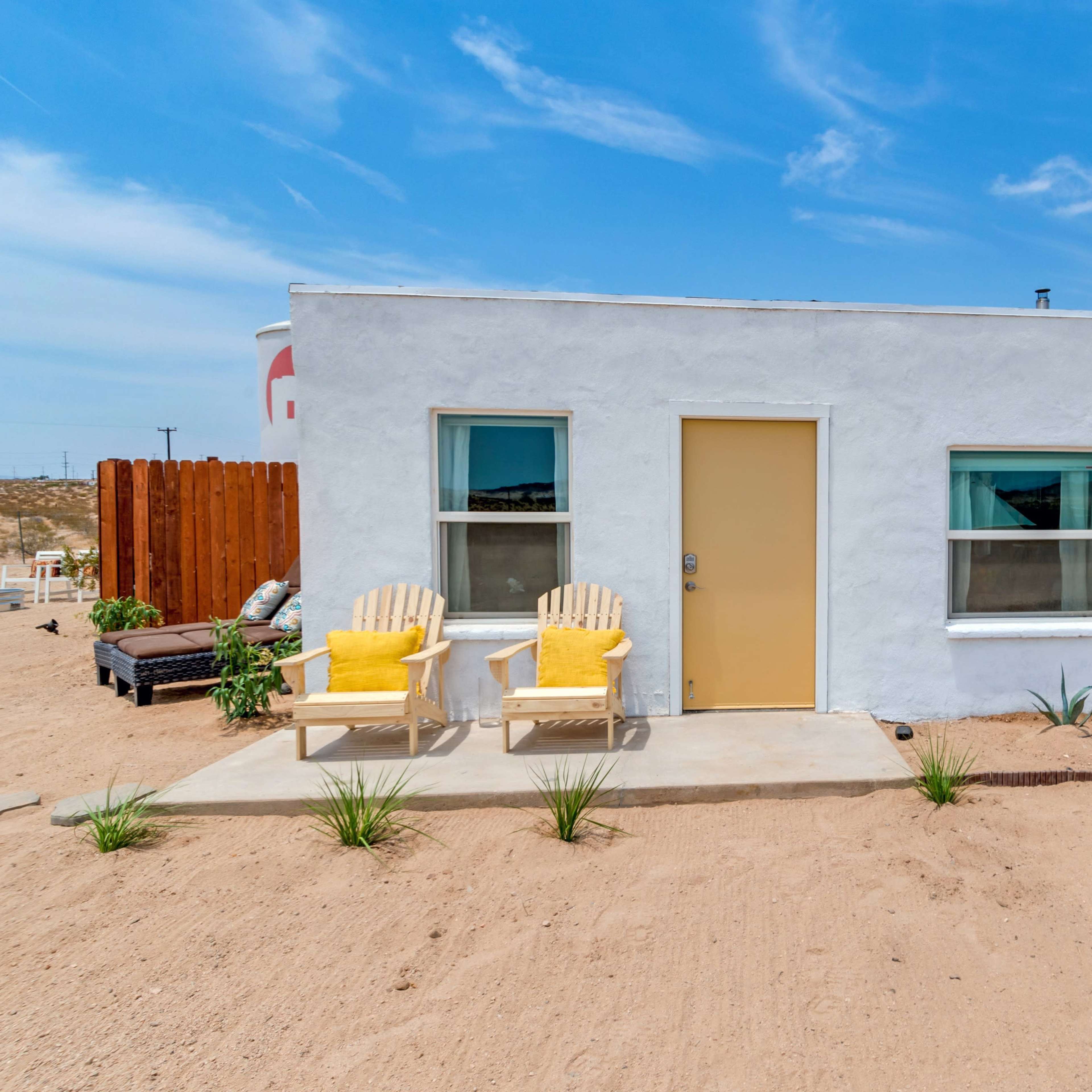 A small white house with a yellow door and two yellow chairs is surrounded by sandy terrain and sparse vegetation.