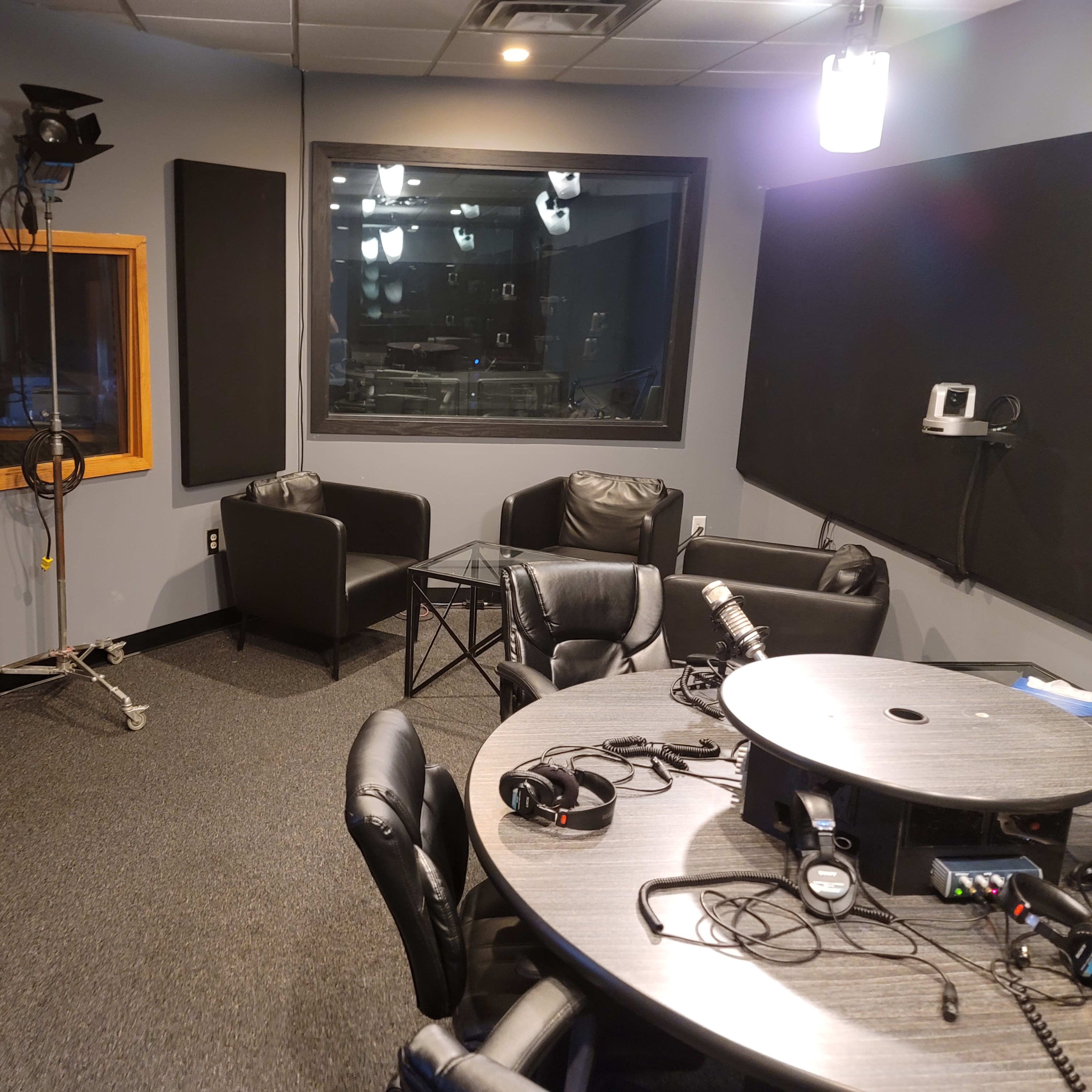 A radio studio with a round table, chairs, and sound equipment, featuring a soundproof glass window.