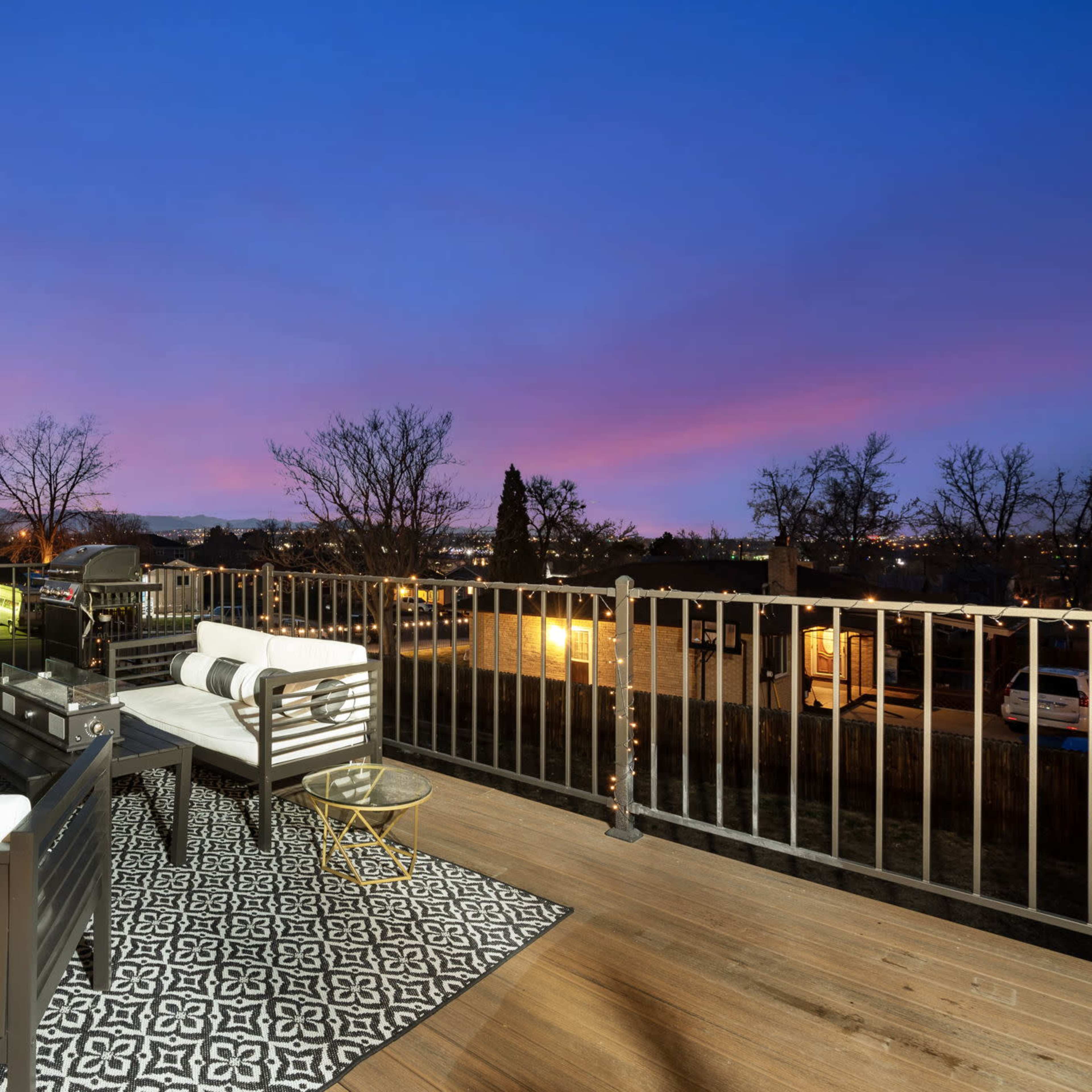 A balcony with modern furniture overlooks a sunset view, featuring a black and white patterned rug and trees in the background.