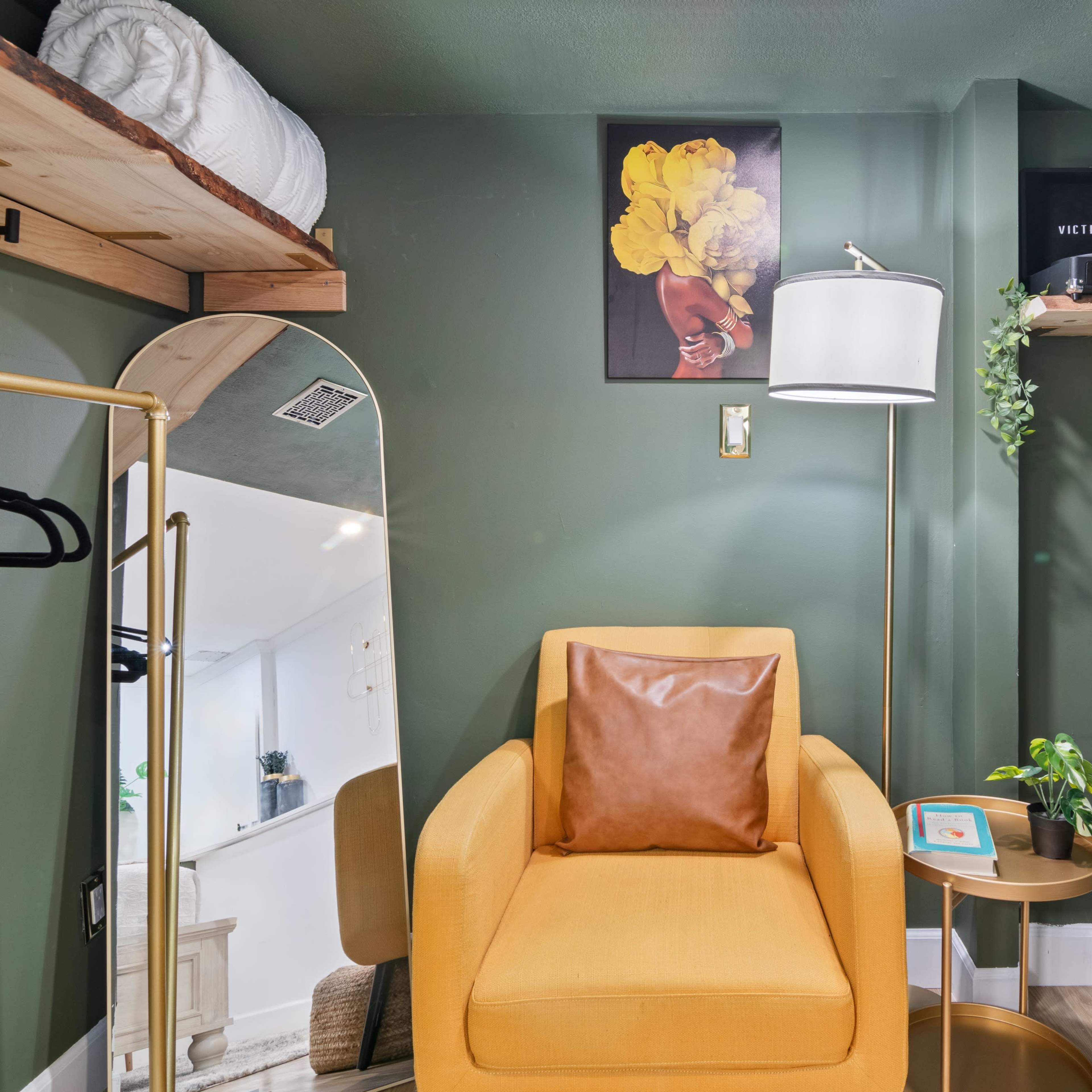 A small corner featuring a yellow armchair, a floor lamp, a mirror, and shelves with decorative items against a deep green wall.