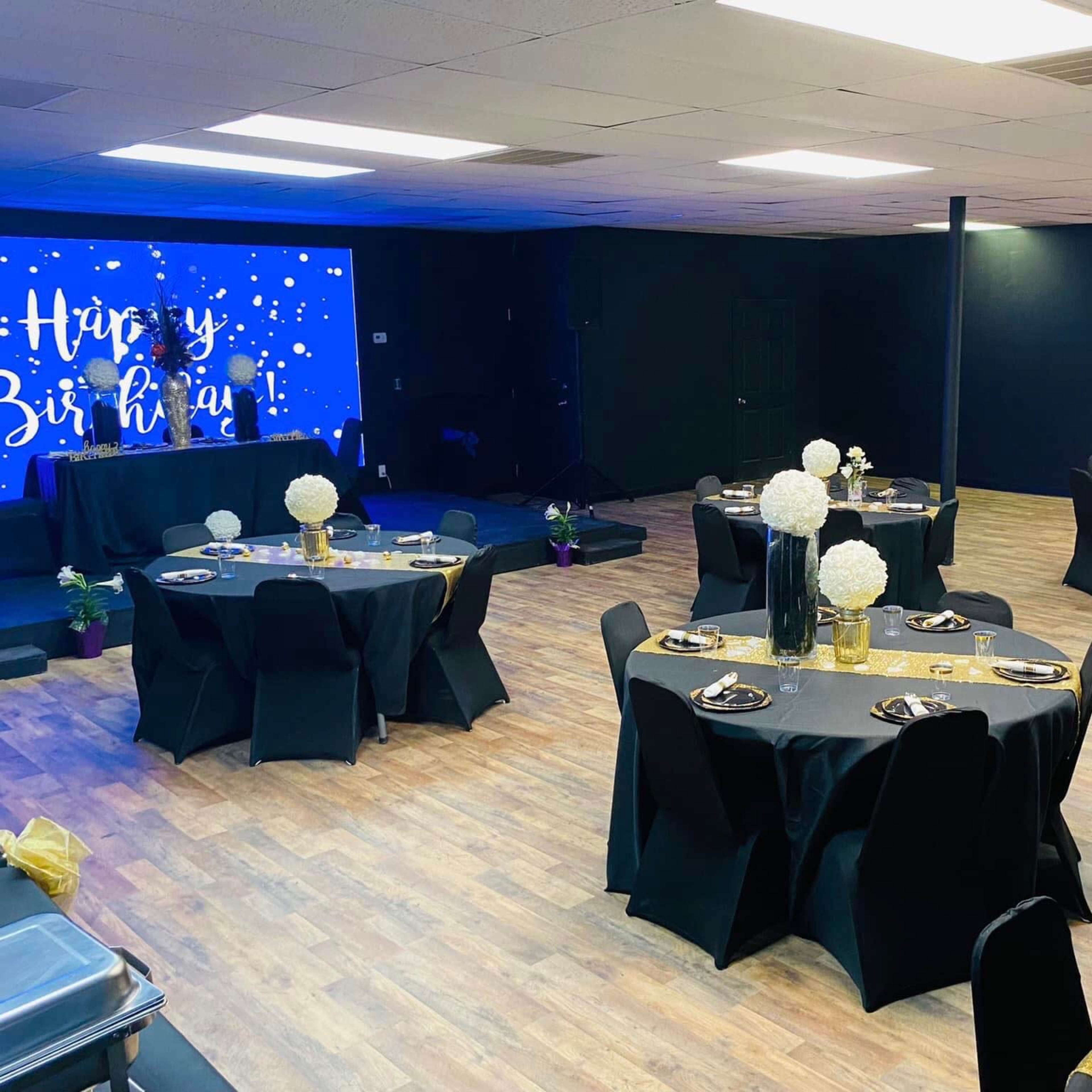 A decorated event space with round tables covered in black tablecloths, each set with floral centerpieces and gold accents, and a backdrop featuring a "Happy Birthday" message.