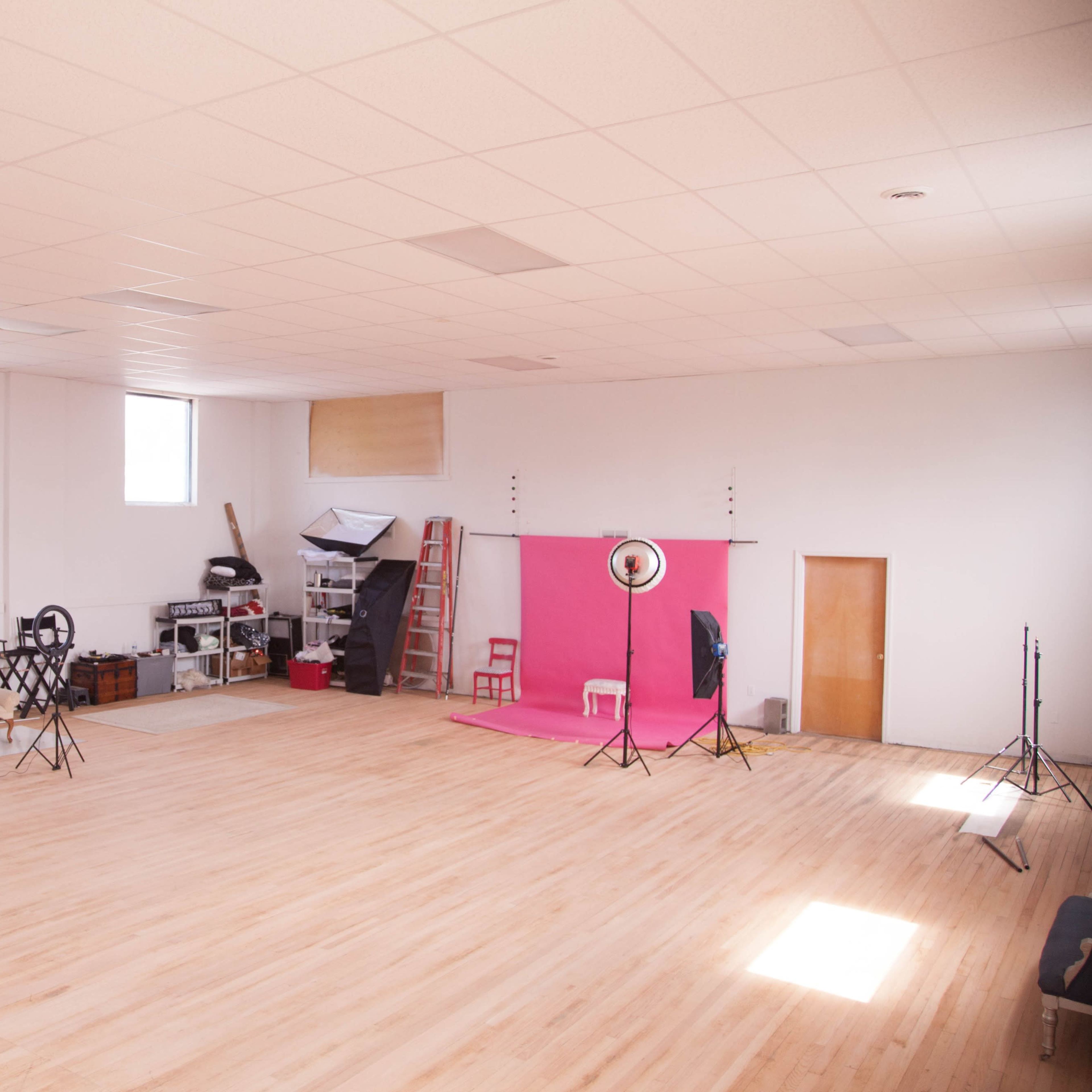 A spacious photography studio features hardwood floors, large windows, and a pink backdrop setup with lighting equipment on one side.