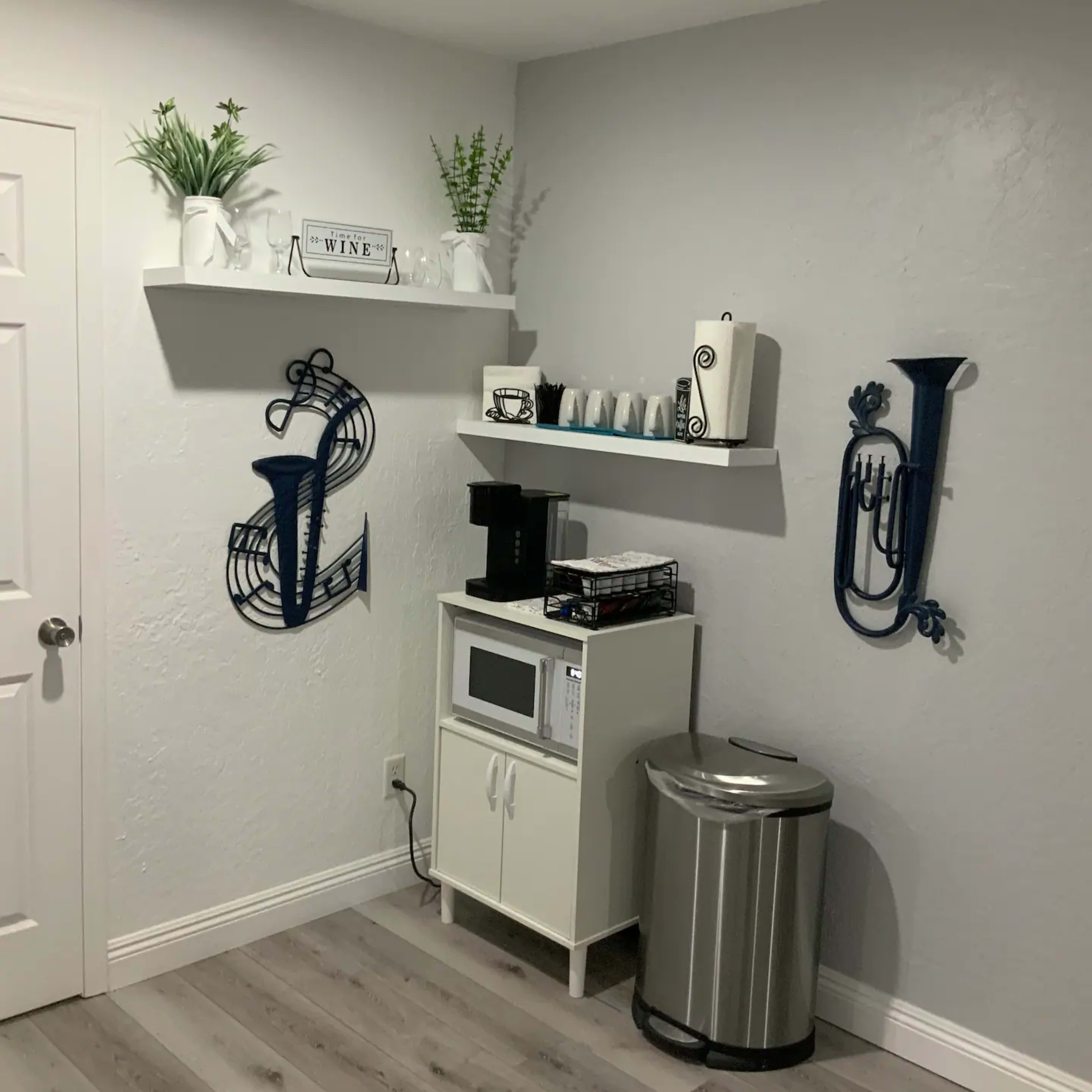 A small kitchen corner featuring a microwave, a coffee maker, a trash can, and decorative shelves with plants and musical instrument wall art.